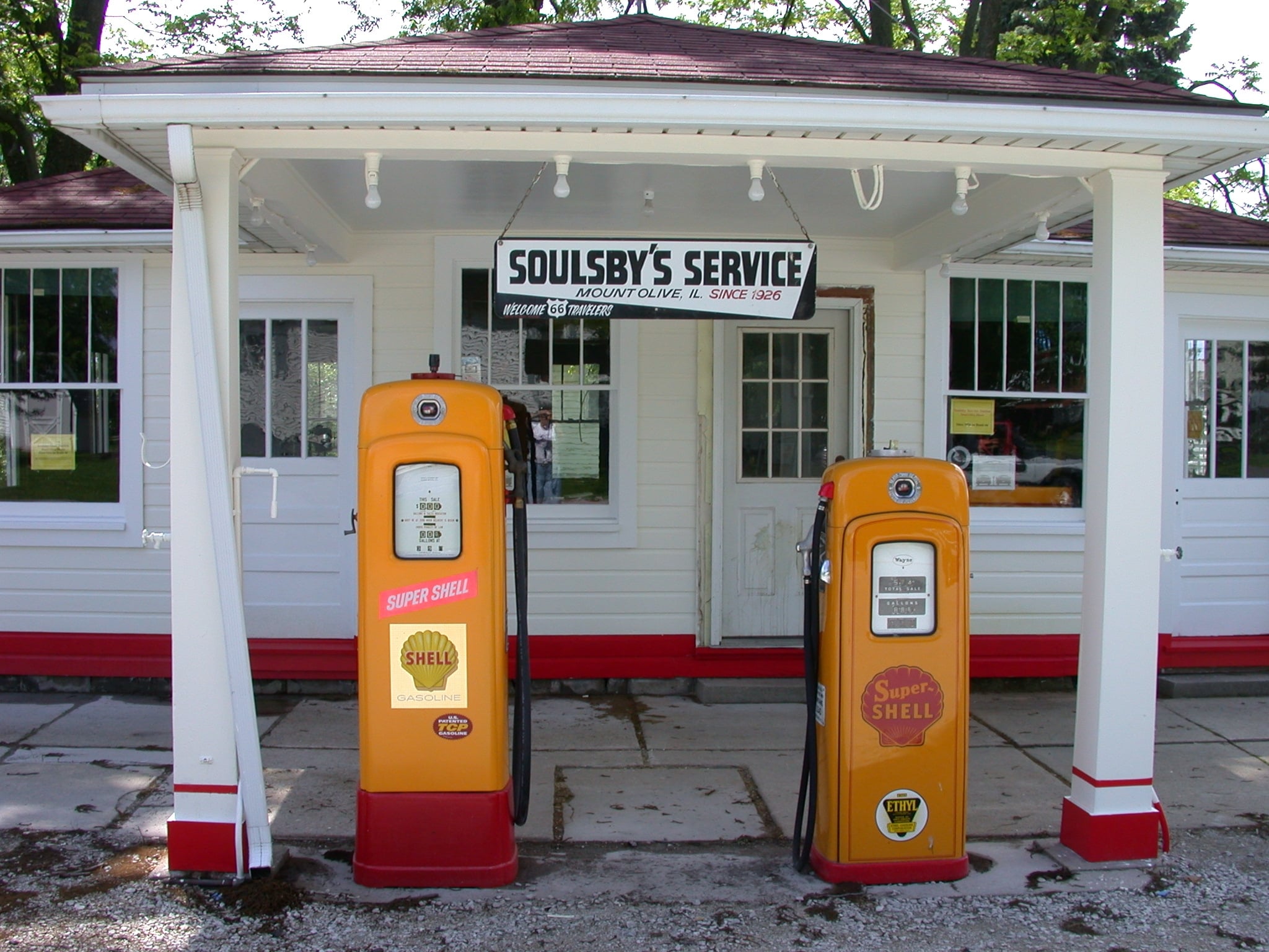 Two vintage yellow and orange Shell gas pumps stand in front of a white building with a sign reading “Soulsby’s Service.” The building has a covered porch, windows, and trees in the background.