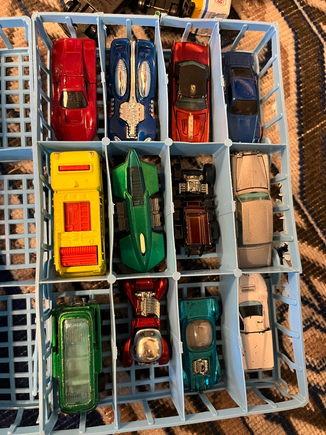 A blue plastic case holds twelve toy cars of various colors and styles, arranged neatly in three rows. The cars include classic, sports, and fantasy models on a patterned fabric surface.