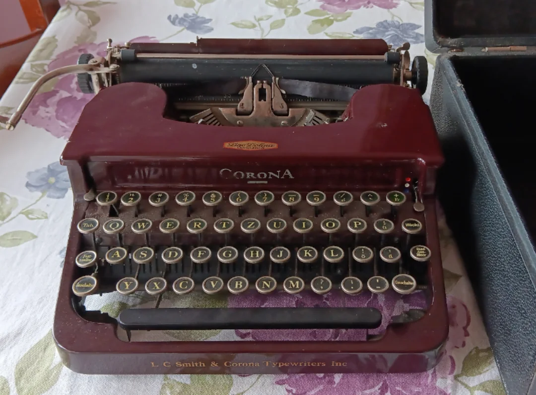 A vintage burgundy Corona typewriter sits on a floral tablecloth, with round black keys and gold lettering. Part of its black carrying case is visible on the right side of the image.