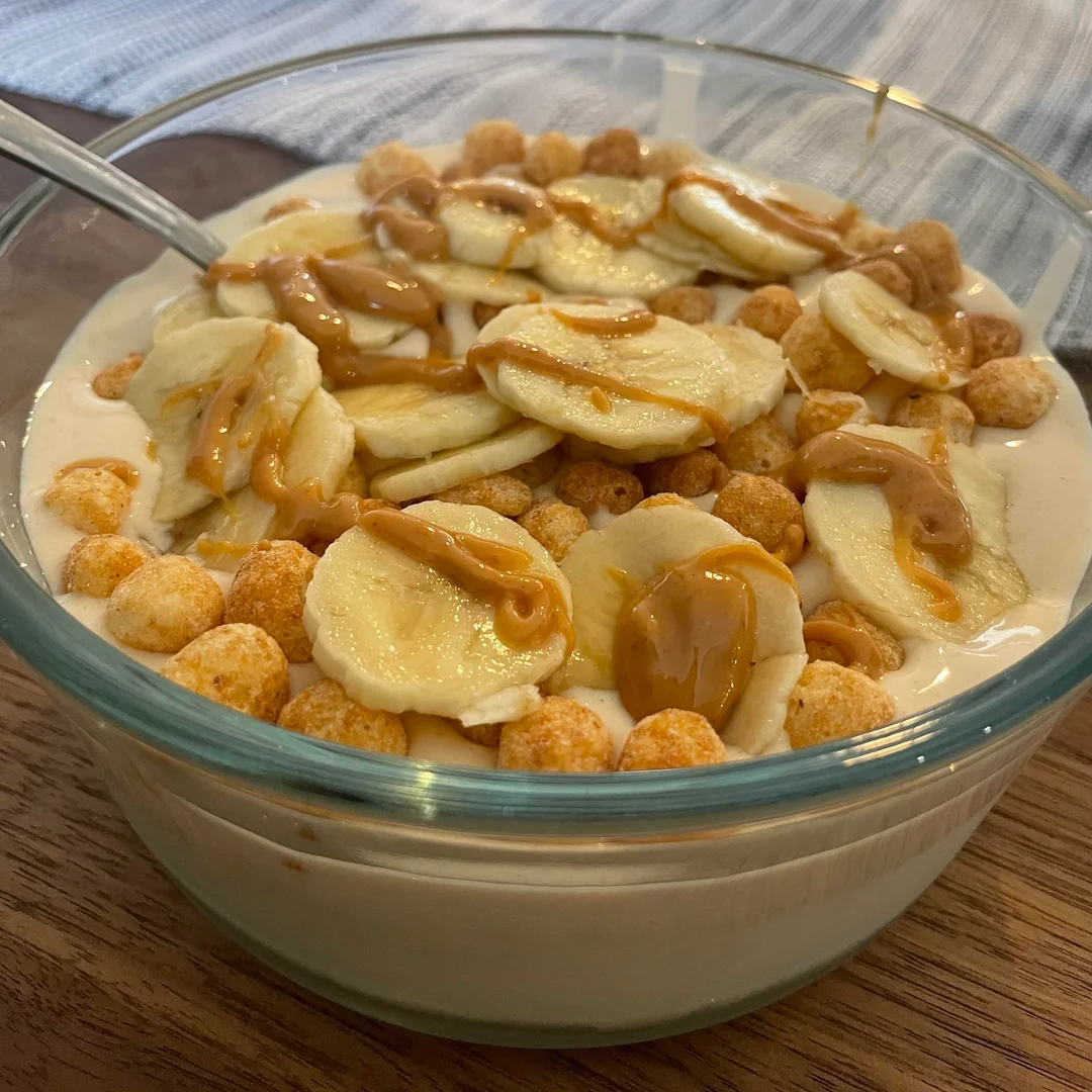A glass bowl filled with yogurt, topped with banana slices, cereal puffs, and drizzled peanut butter. A spoon rests in the bowl.