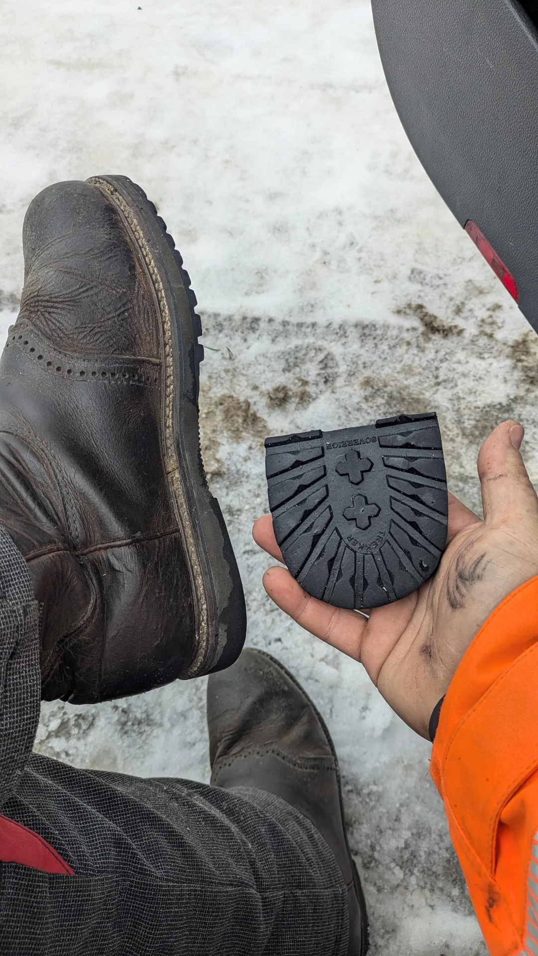 A person holds a black rubber shoe heel in one hand, showing the detached sole. Their other foot wears a brown, worn leather shoe. The ground is snowy and dirty.
