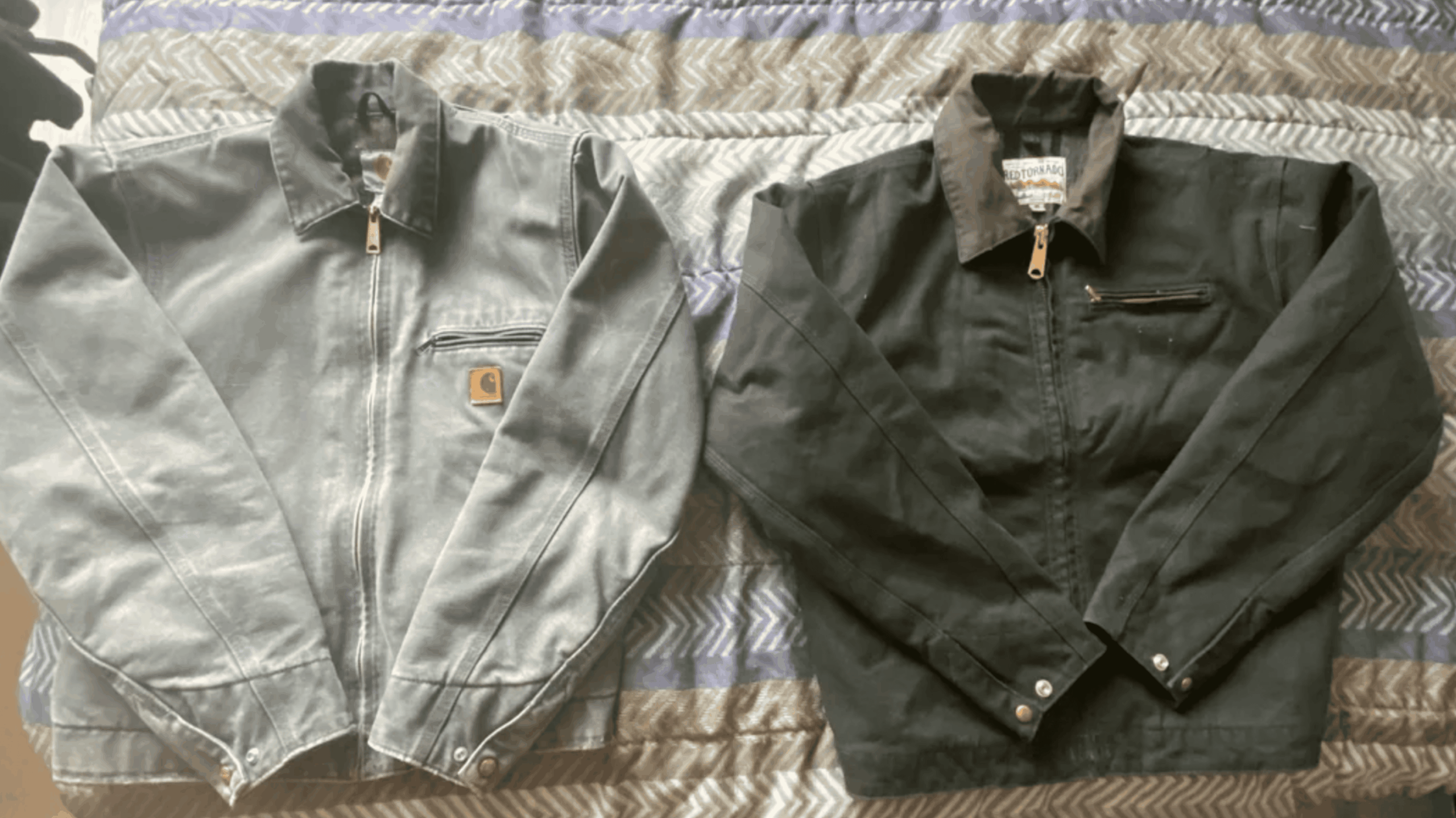 Two work jackets, one gray and one black, are laid flat side by side on a patterned surface. Both have zippered chest pockets, collars, and metal buttons on the cuffs.