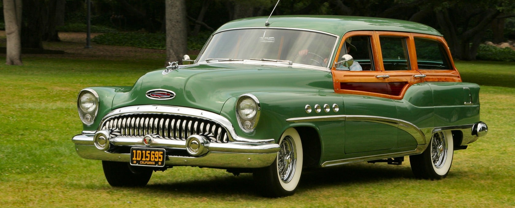 A green vintage station wagon with wood paneling is parked on grass in a park-like setting, featuring whitewall tires and classic chrome details.