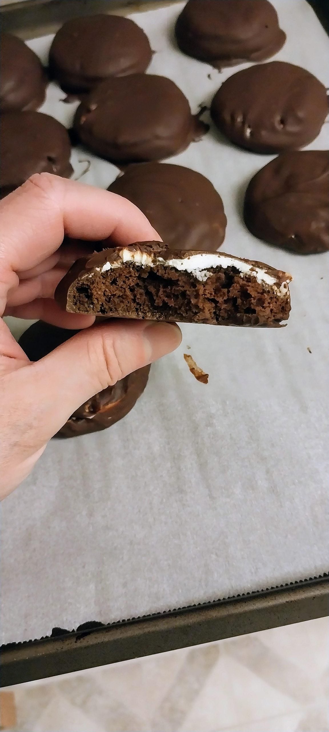 A hand holds a half-eaten chocolate cookie with a white filling. More whole chocolate-covered cookies are on a parchment-lined baking tray in the background.