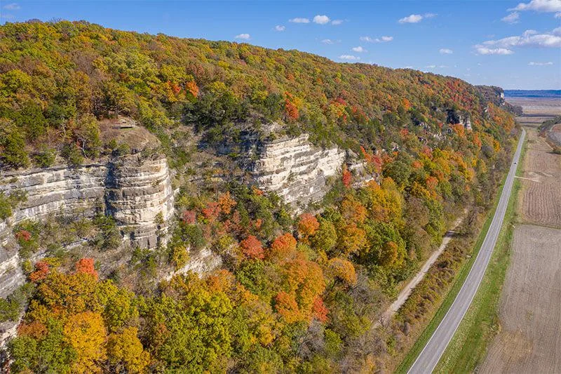 A scenic aerial view of a forested limestone bluff with autumn foliage, bordering a rural road and open farmland under a blue sky with scattered clouds.
