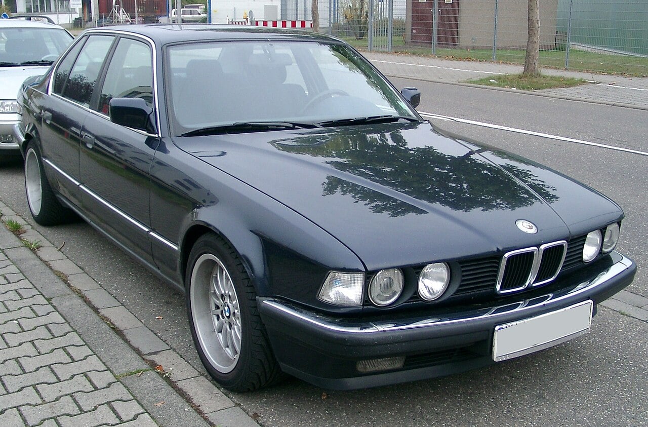 A dark blue BMW sedan is parked on the street near a curb. The car features classic round headlights and alloy wheels, with trees and buildings visible in the background.