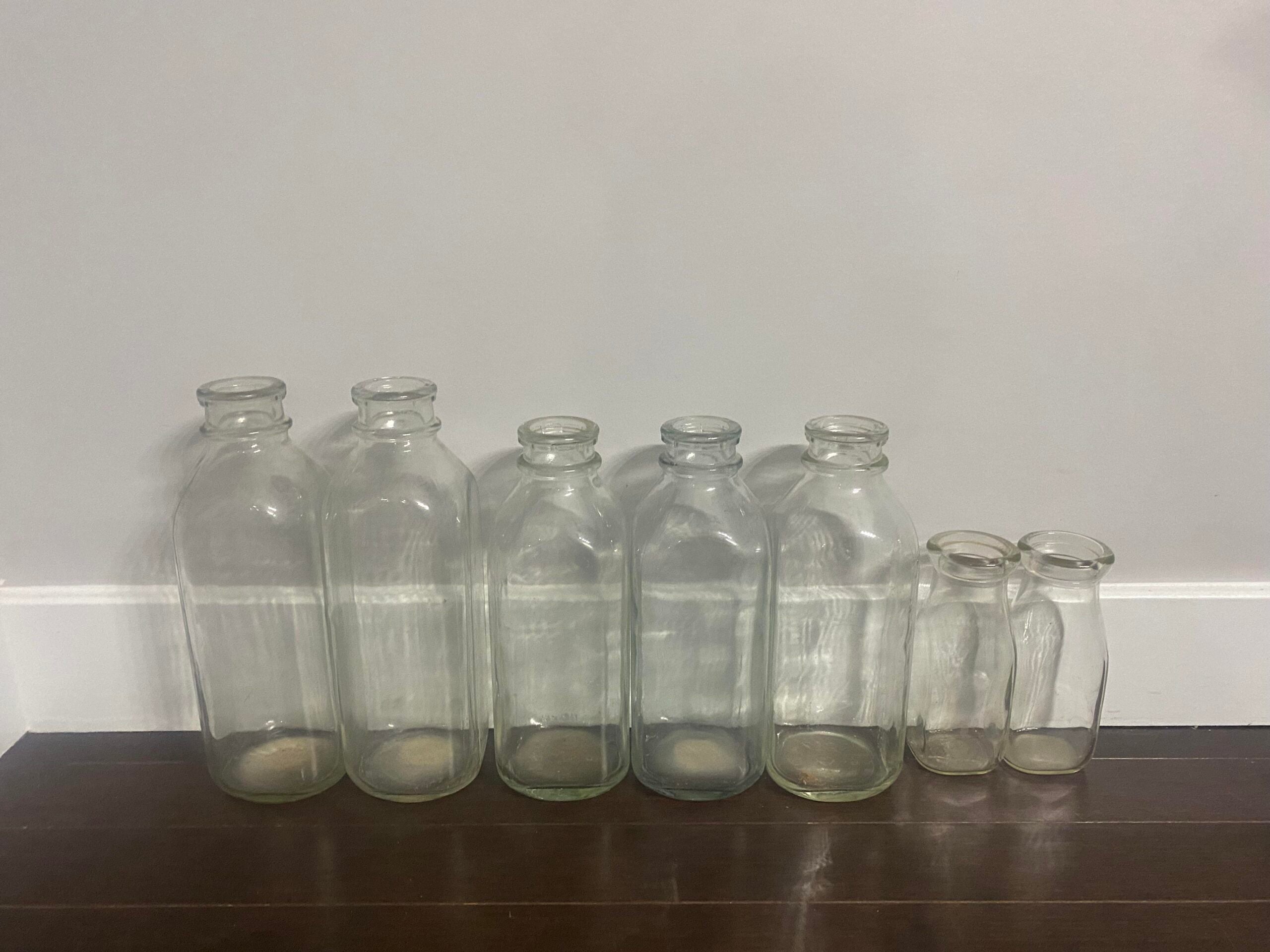Seven empty glass bottles of varying sizes are lined up on a dark wooden floor against a plain light-colored wall.