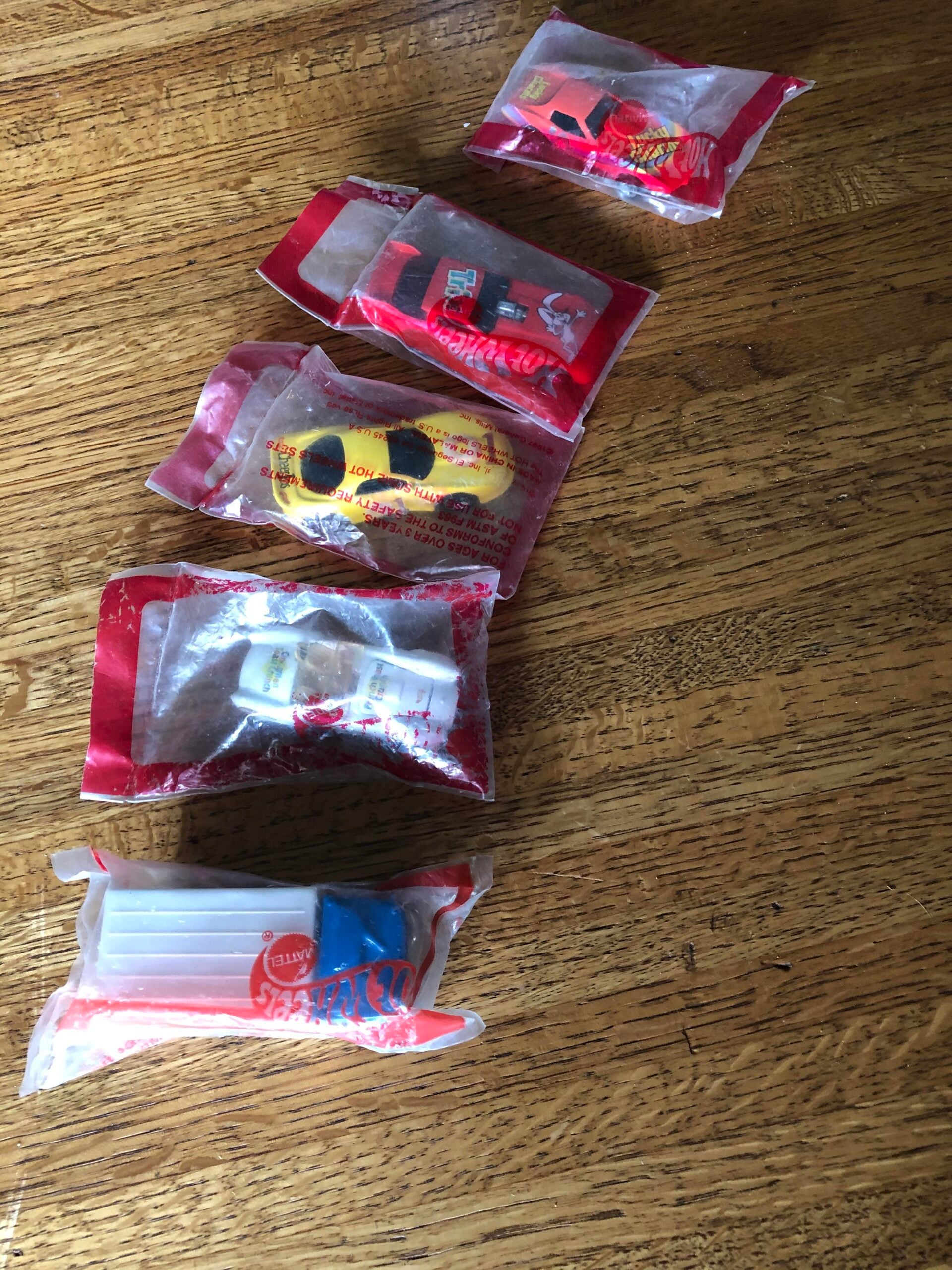 Five small toys in clear plastic bags, each with red and white trim, are arranged in a line on a wooden surface. The toys include cars and other vehicles, each individually packaged.