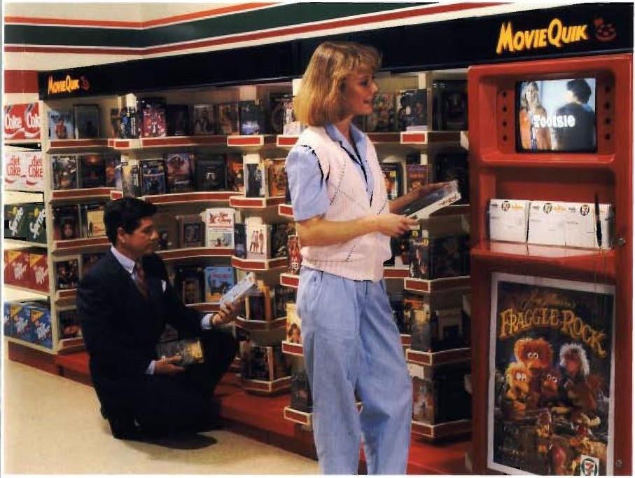A woman in light blue and pink shops for VHS tapes at a 1980s video rental section. A man kneels nearby browsing tapes. Shelves display movies; a screen shows previews; a “Fraggle Rock” tape is visible in front.