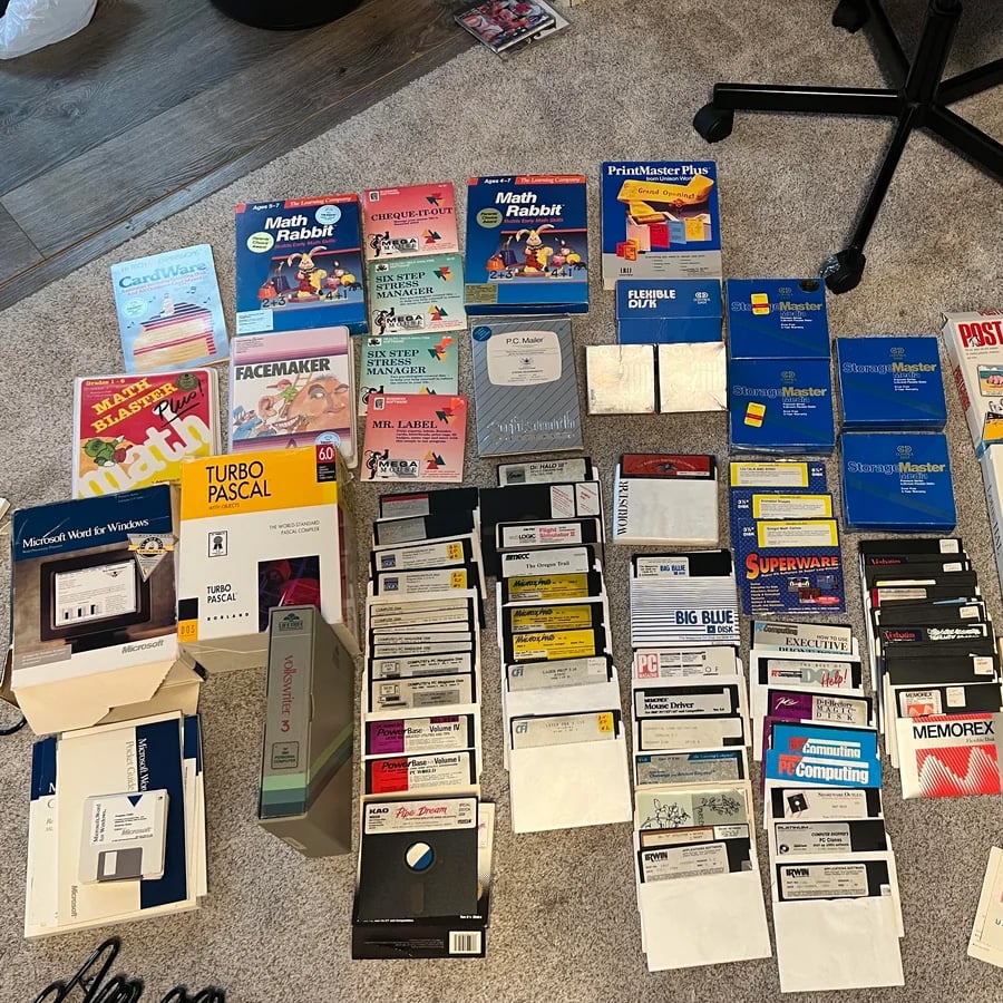 A large collection of vintage floppy disks, manuals, and software boxes are spread out on a carpeted floor, including titles like Math Rabbit, Turbo Pascal, PrintMaster Plus, and various business and educational programs.