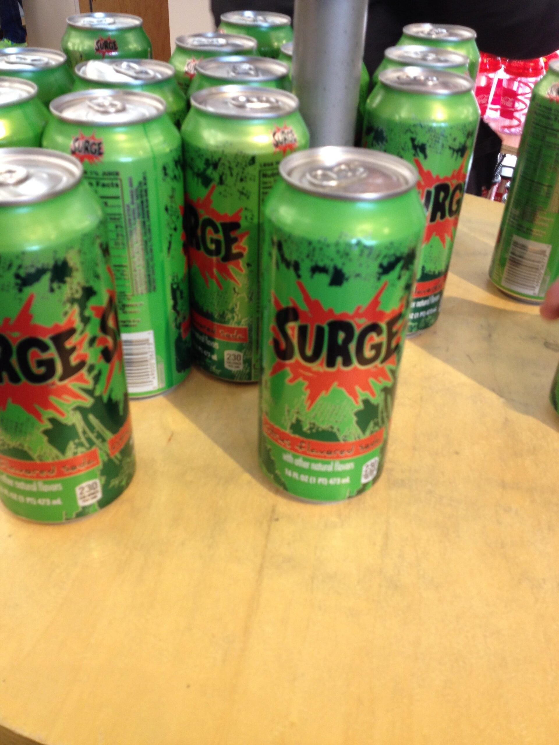 Several green Surge soda cans with red and black graphics are displayed on a light wood surface. One can is prominently in front, while others are grouped together in the background.