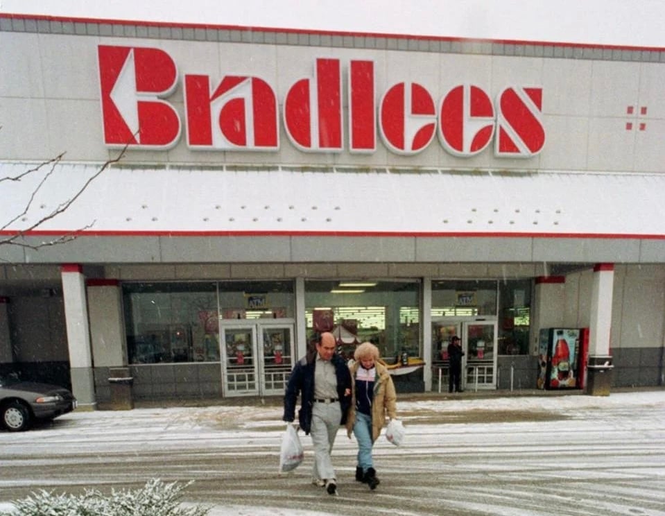 Two people carrying shopping bags walk out of a Bradlees store on a snowy day, with snow covering the parking lot and cars visible on the left.