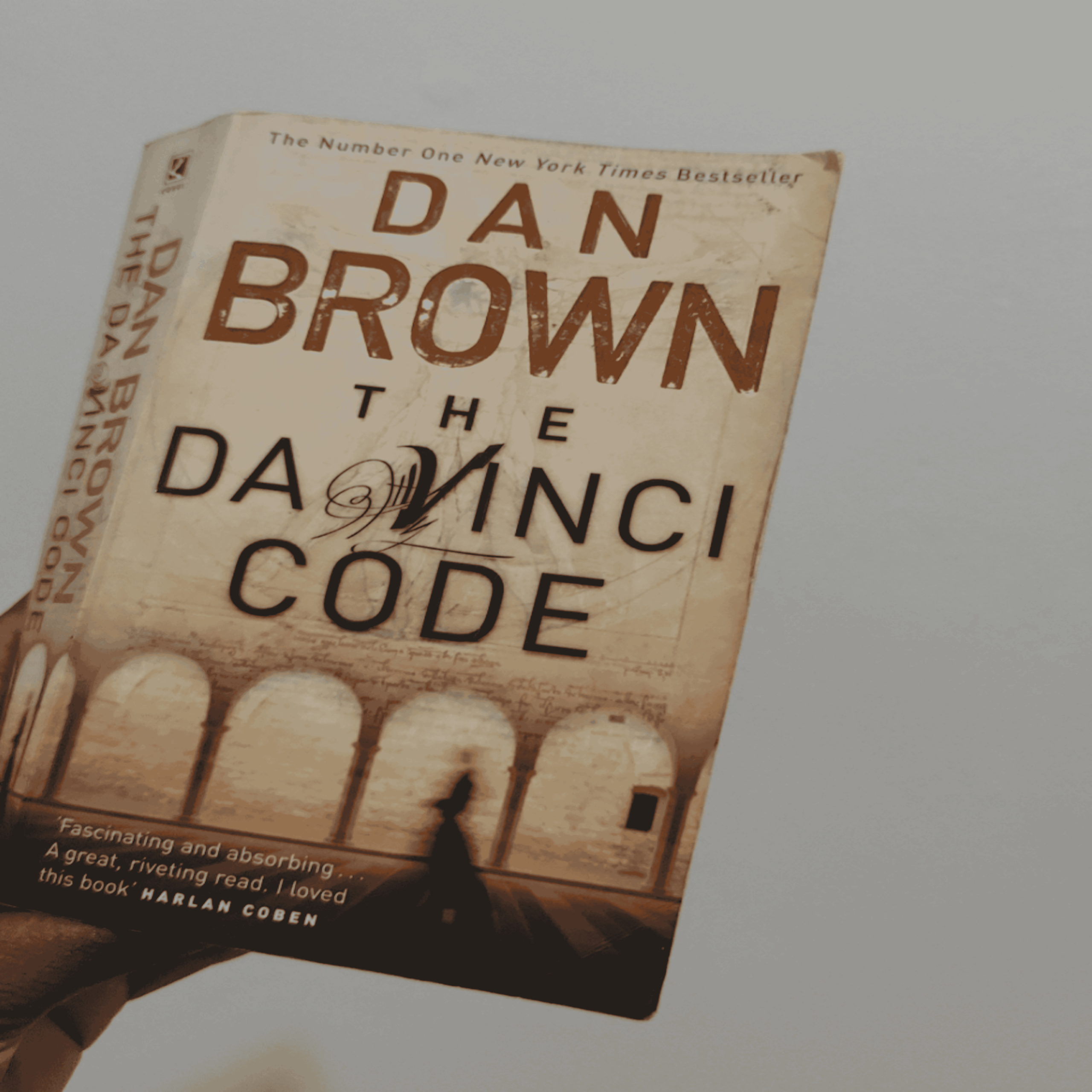 A hand holds a paperback copy of “The Da Vinci Code” by Dan Brown. The cover features arches and a blurred shadowy figure against a beige background with brown text.