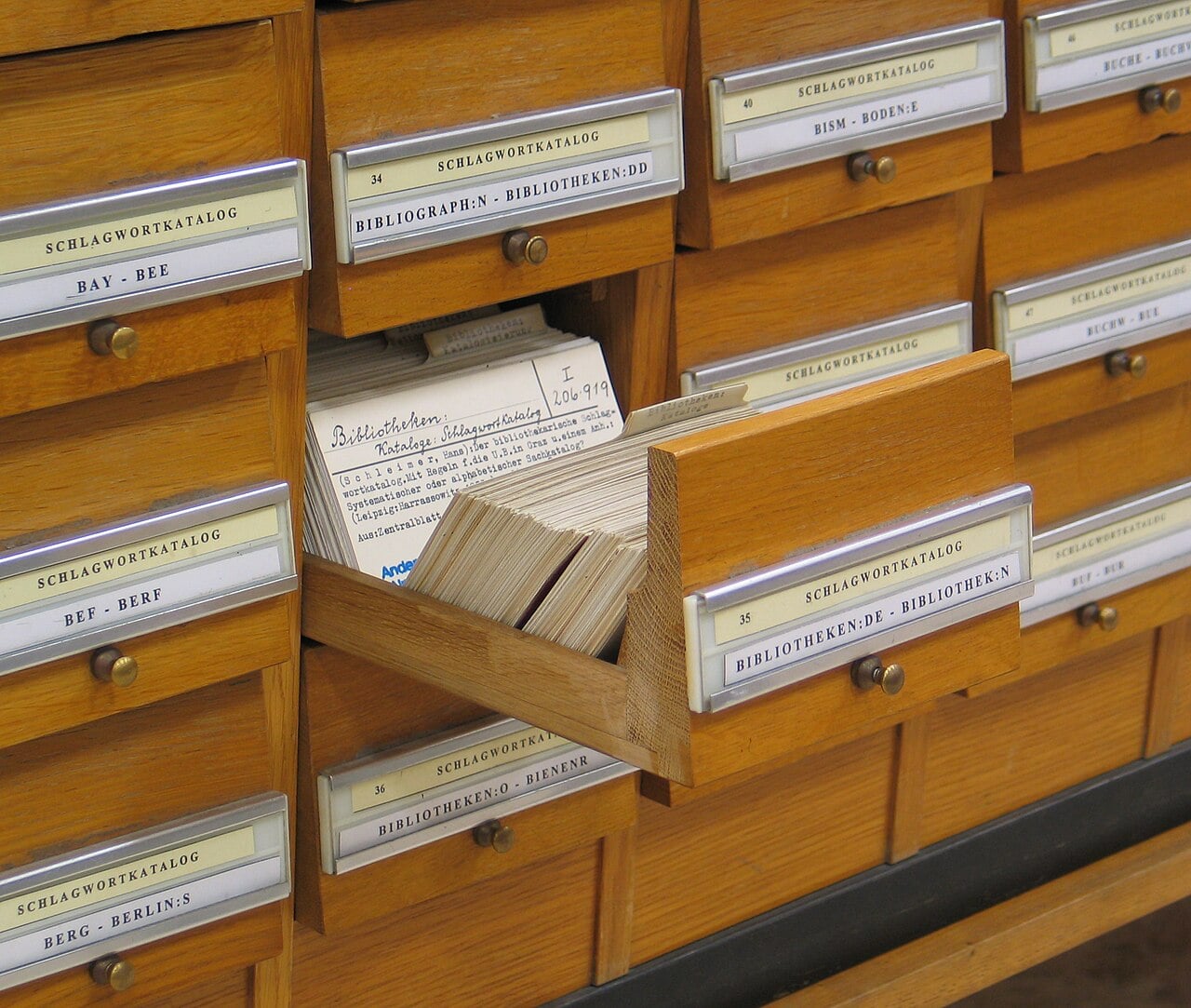 A wooden card catalog with labeled drawers, one drawer open showing index cards with handwritten and typed text, used for organizing library information.