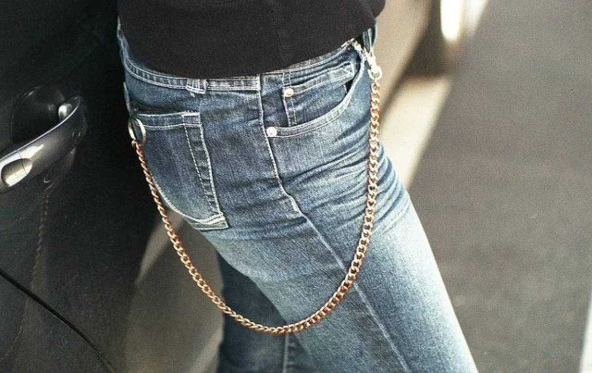 Person wearing blue jeans with a gold chain attached, leaning against a black car. The image focuses on the lower torso and upper legs, not showing the person’s face.