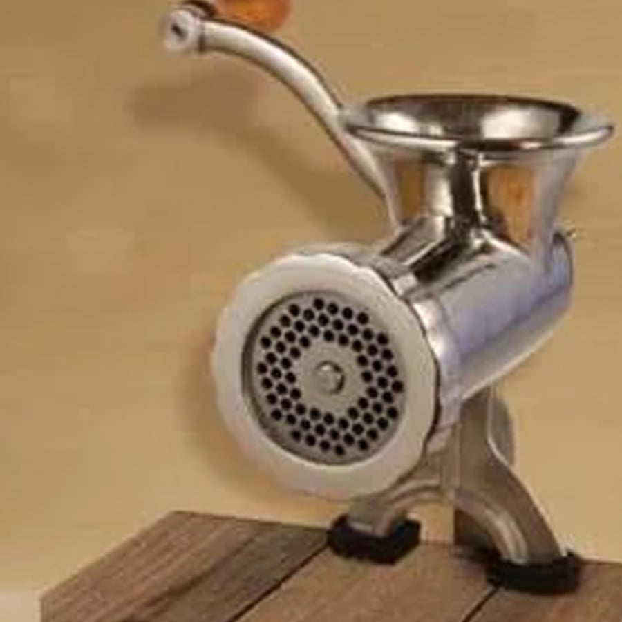 A metal manual meat grinder with a wooden handle is clamped to a wooden surface, against a plain beige background.