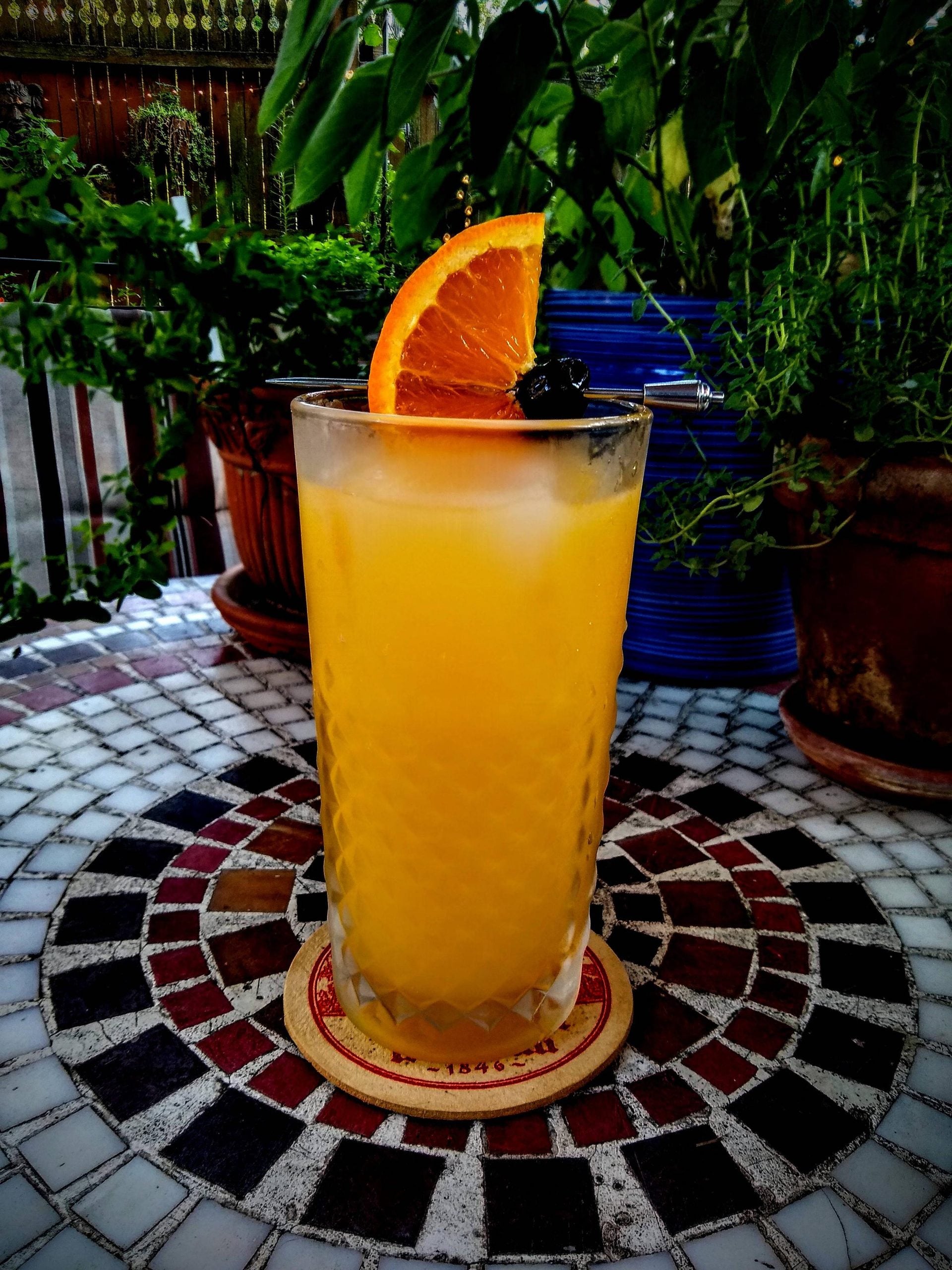 A tall glass of orange juice garnished with an orange slice and a cherry sits on a coaster on a mosaic table, surrounded by potted plants.