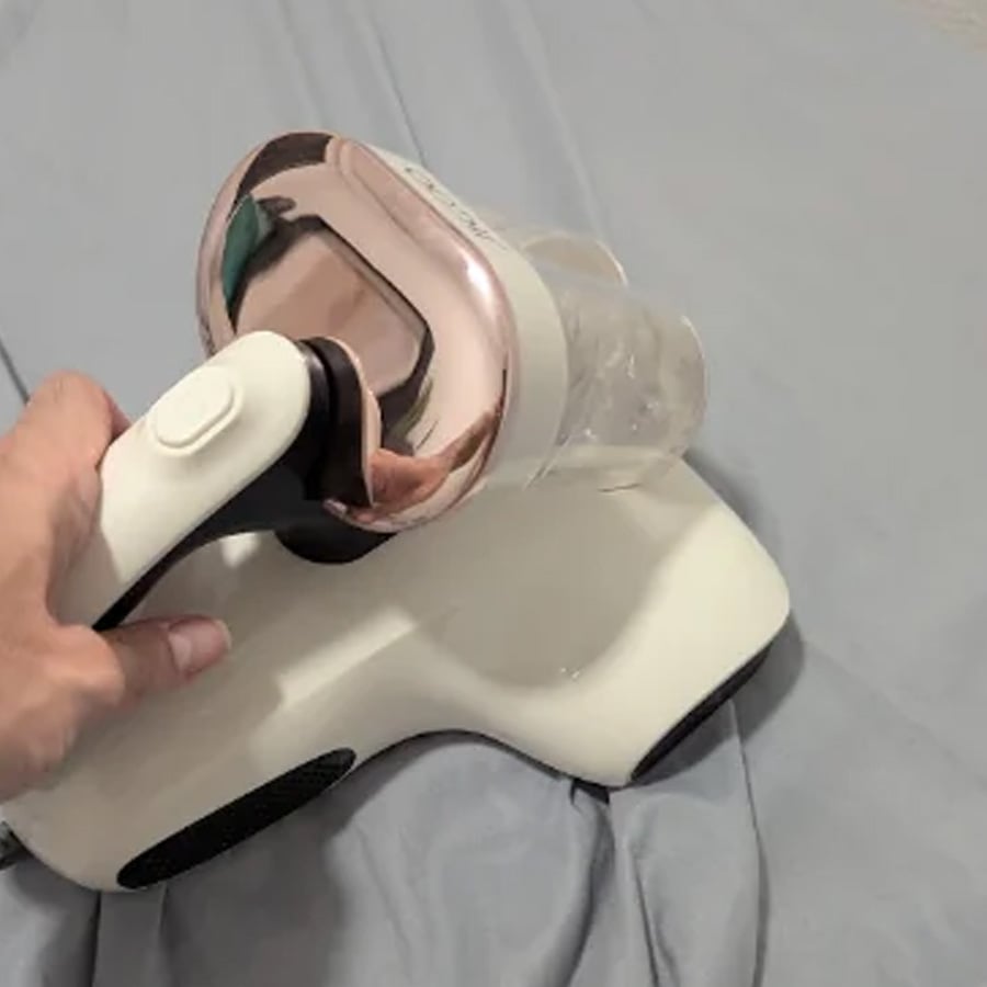 A hand is holding a white and rose gold handheld vacuum cleaner, using it on a light blue fabric surface, possibly a bedsheet.