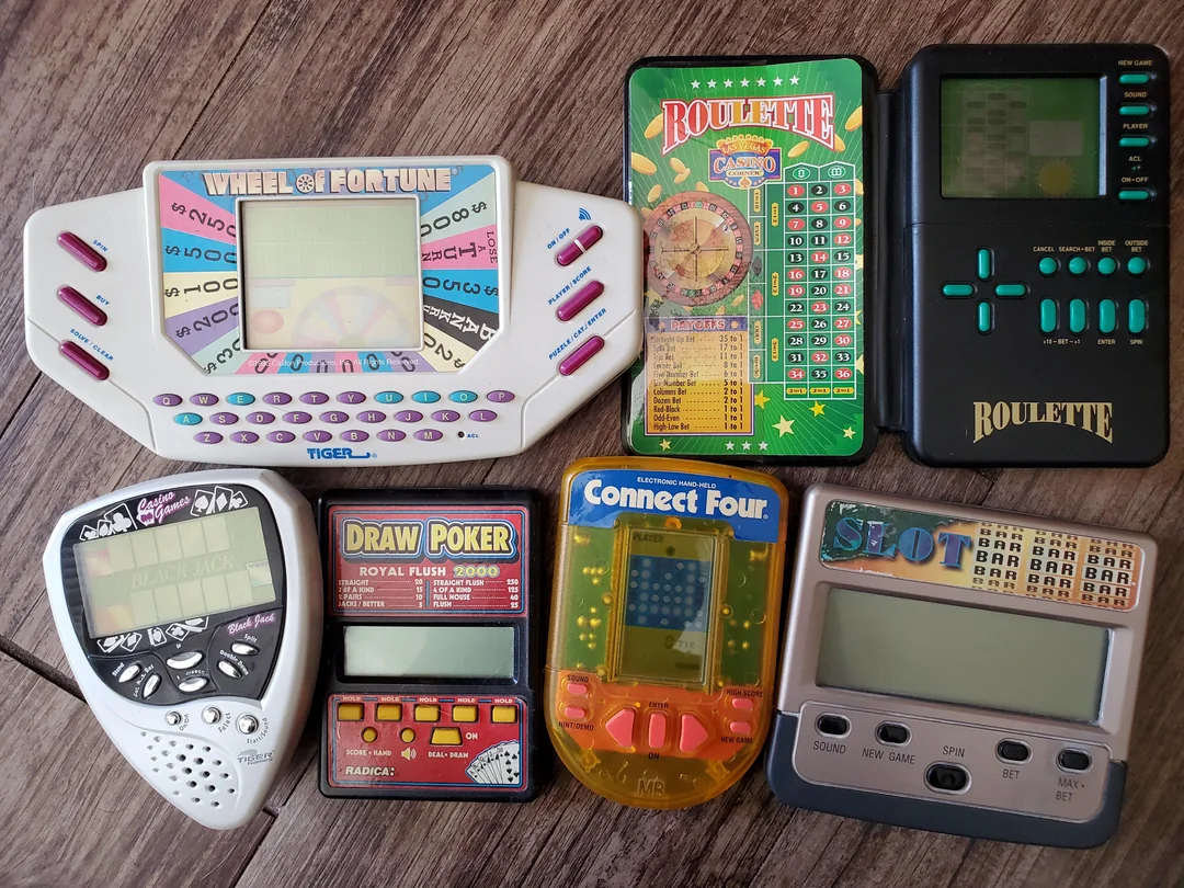 Seven vintage handheld electronic games, including Wheel of Fortune, Roulette, Draw Poker, Connect Four, and Slot, are arranged on a wooden surface. Each device has distinctive colors, buttons, and display screens.