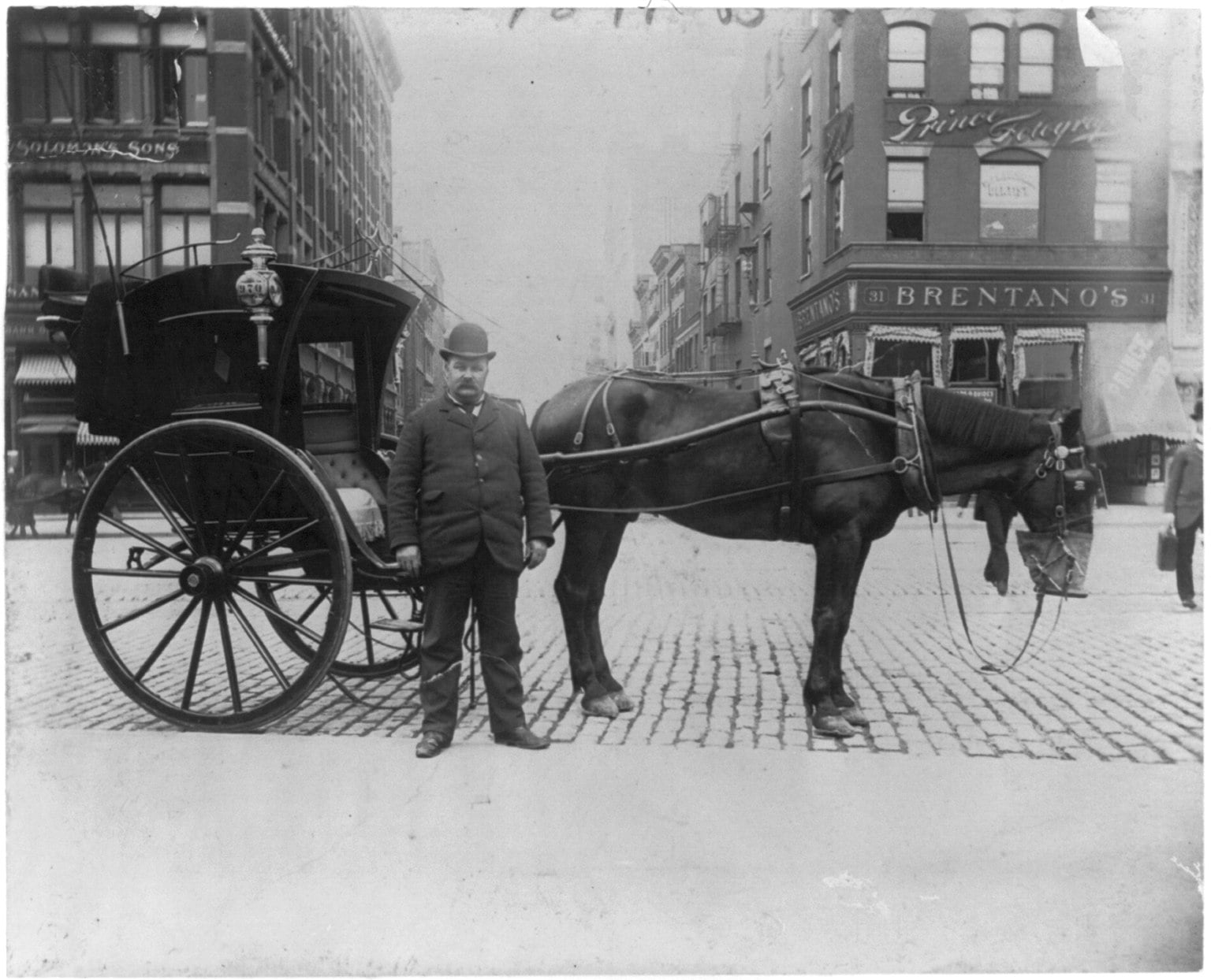 A man in a suit and bowler hat stands beside a horse-drawn carriage on a cobblestone city street, with brick buildings and shop signs visible in the background.