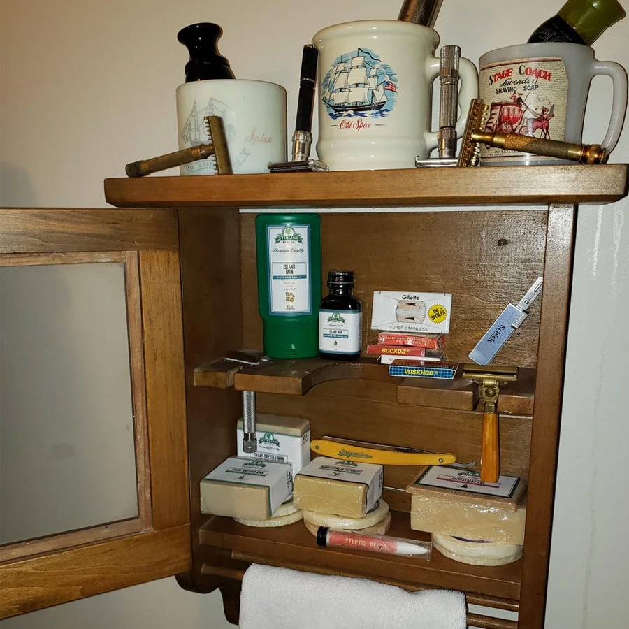 Wooden bathroom cabinet with mugs, shaving brushes, razors, soap bars, aftershave bottles, and shaving accessories organized on shelves. A towel hangs below the cabinet.