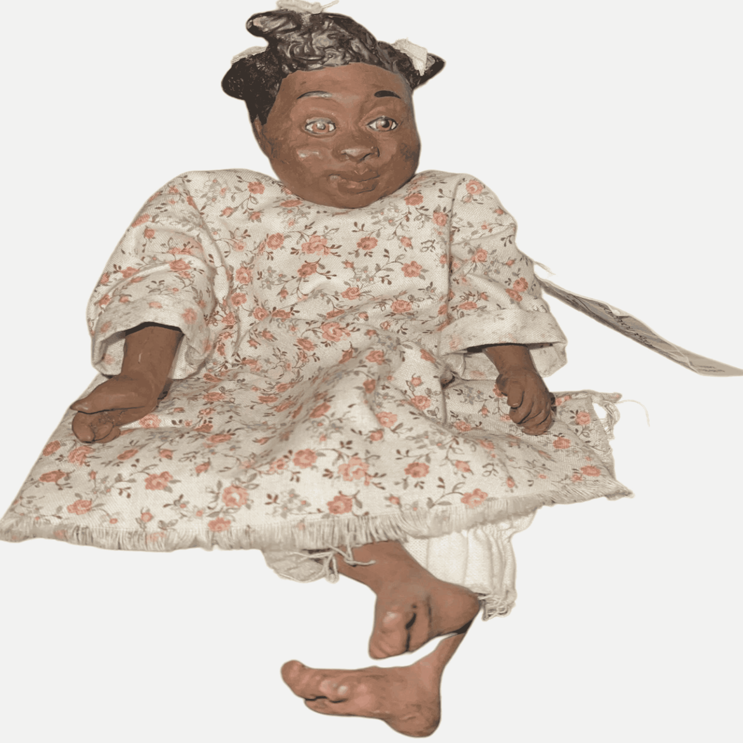 A handmade doll with dark skin, black hair tied with a ribbon, and wearing a floral dress and white bloomers, sitting with bare feet showing.