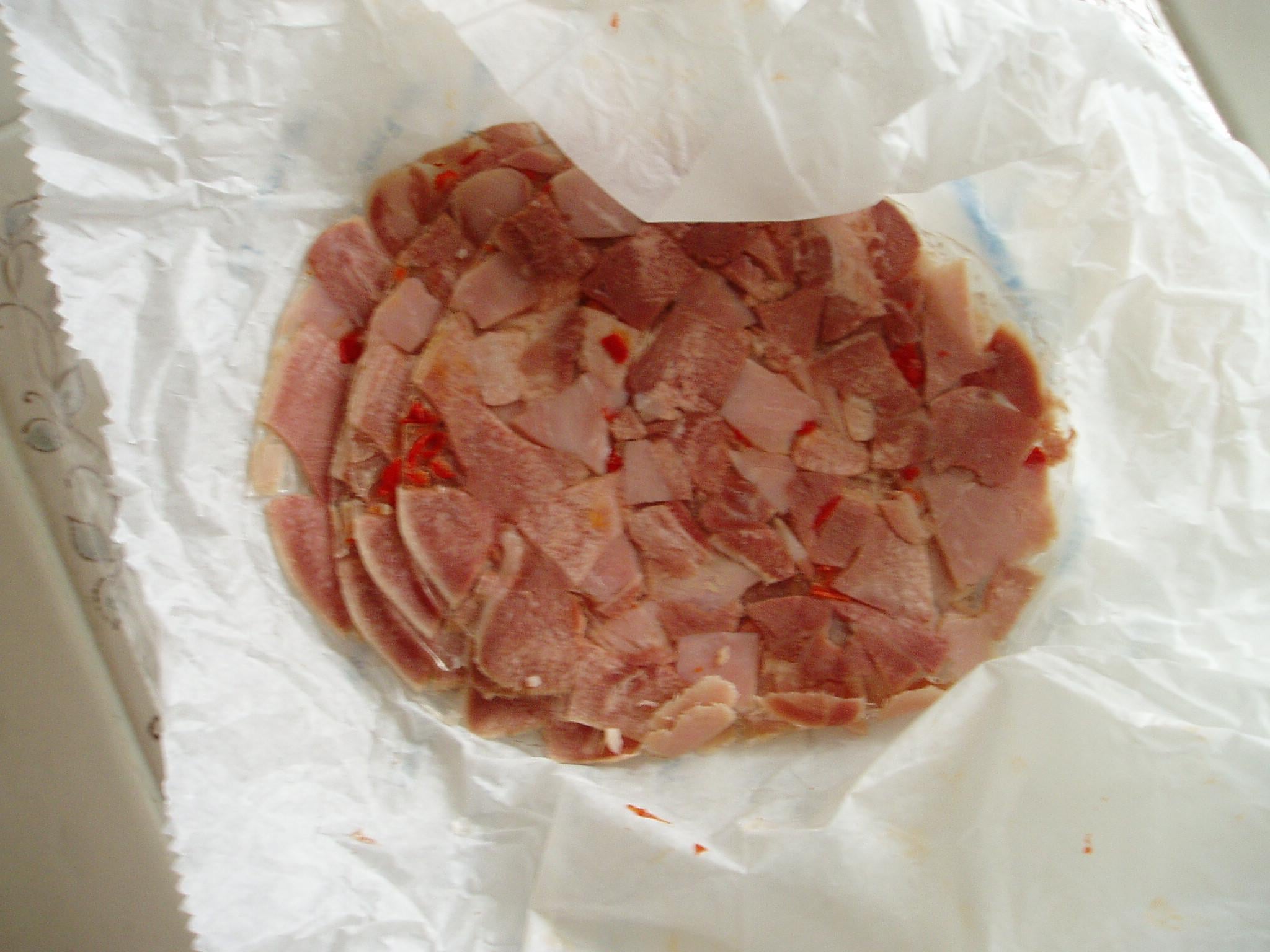 A round slice of luncheon meat with visible chunks and red pepper pieces sits on crumpled white paper, viewed from above.