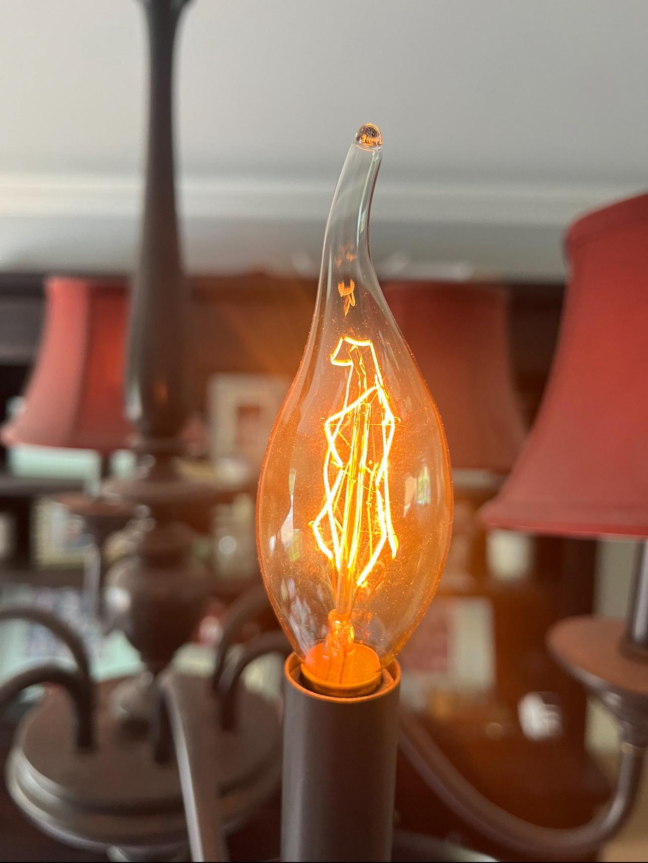 A close-up of a lit candle-shaped light bulb with a glowing filament, surrounded by red lampshades and dark metal lamp arms in the background.