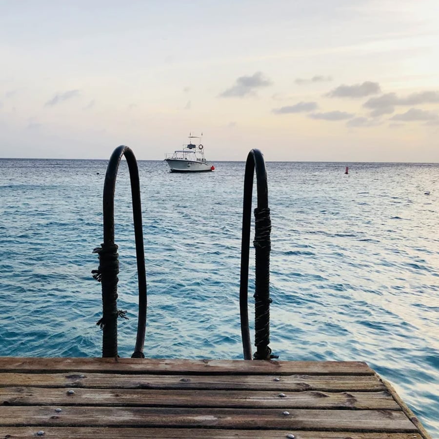 Metal ladder on a wooden dock leading into calm blue ocean water, with a white boat floating in the distance under a cloudy sky at sunset.