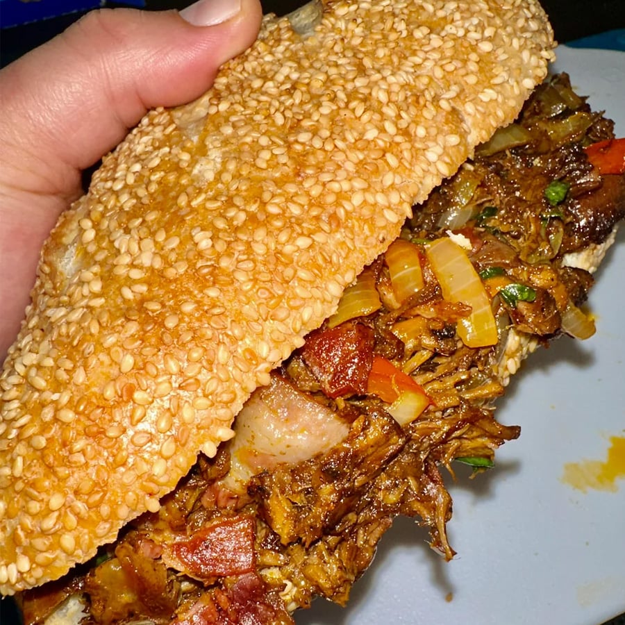 A hand holds a sesame seed sandwich filled with seasoned shredded meat, onions, and diced vegetables.
