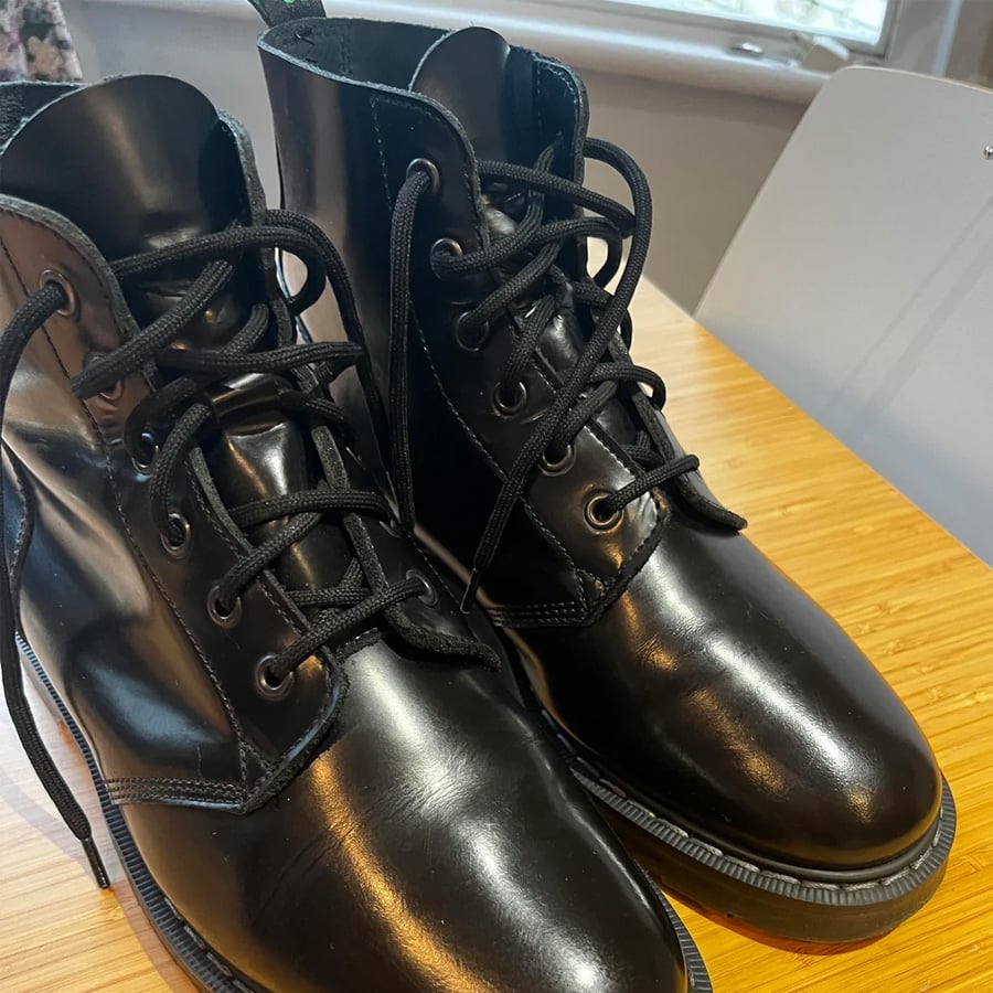 A pair of shiny black lace-up boots sits on a wooden table next to a light grey chair, with a window and some greenery visible in the background.