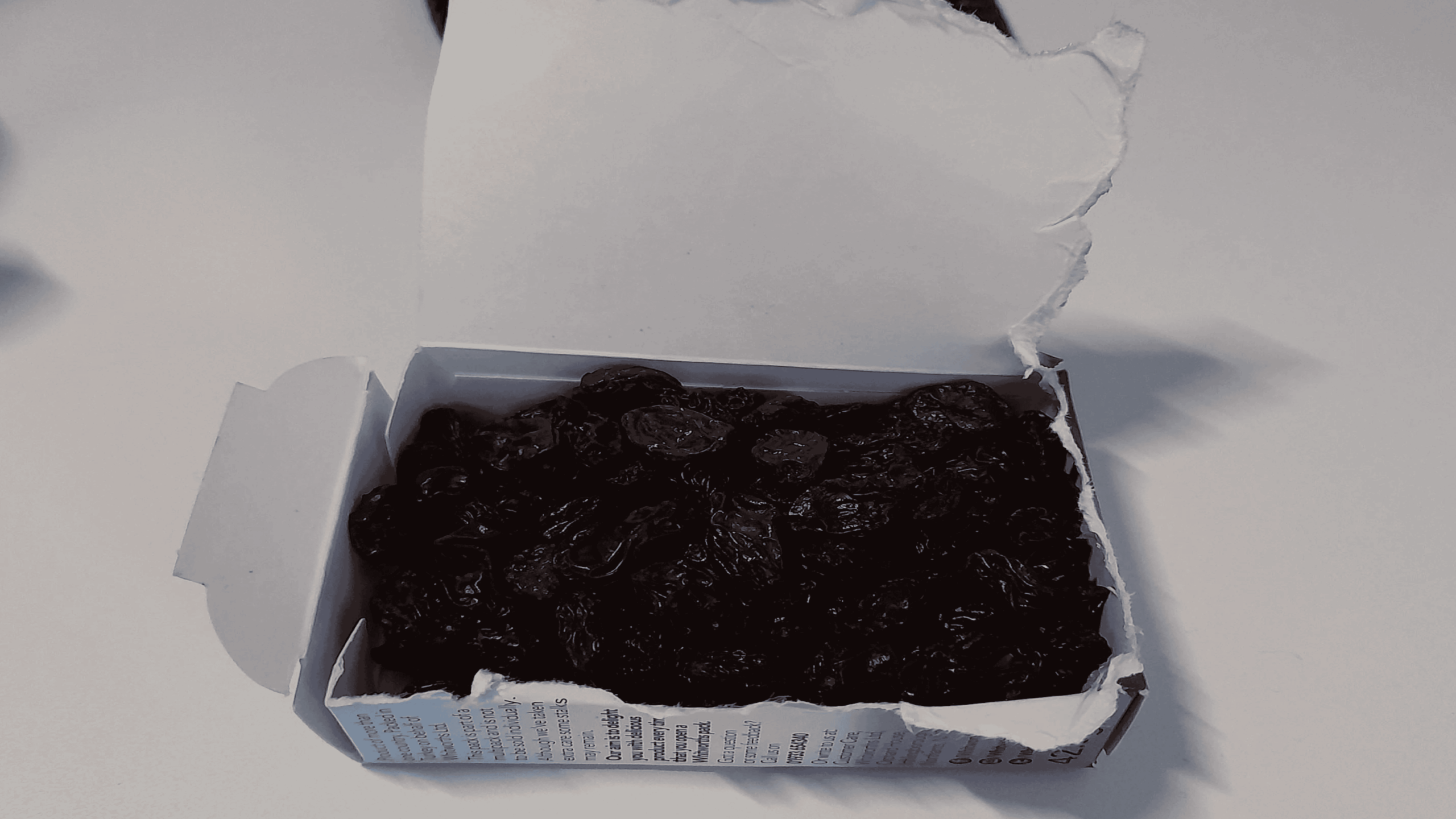 An open cardboard box filled with raisins sits on a white surface, with its lid partially torn back to reveal the contents inside.