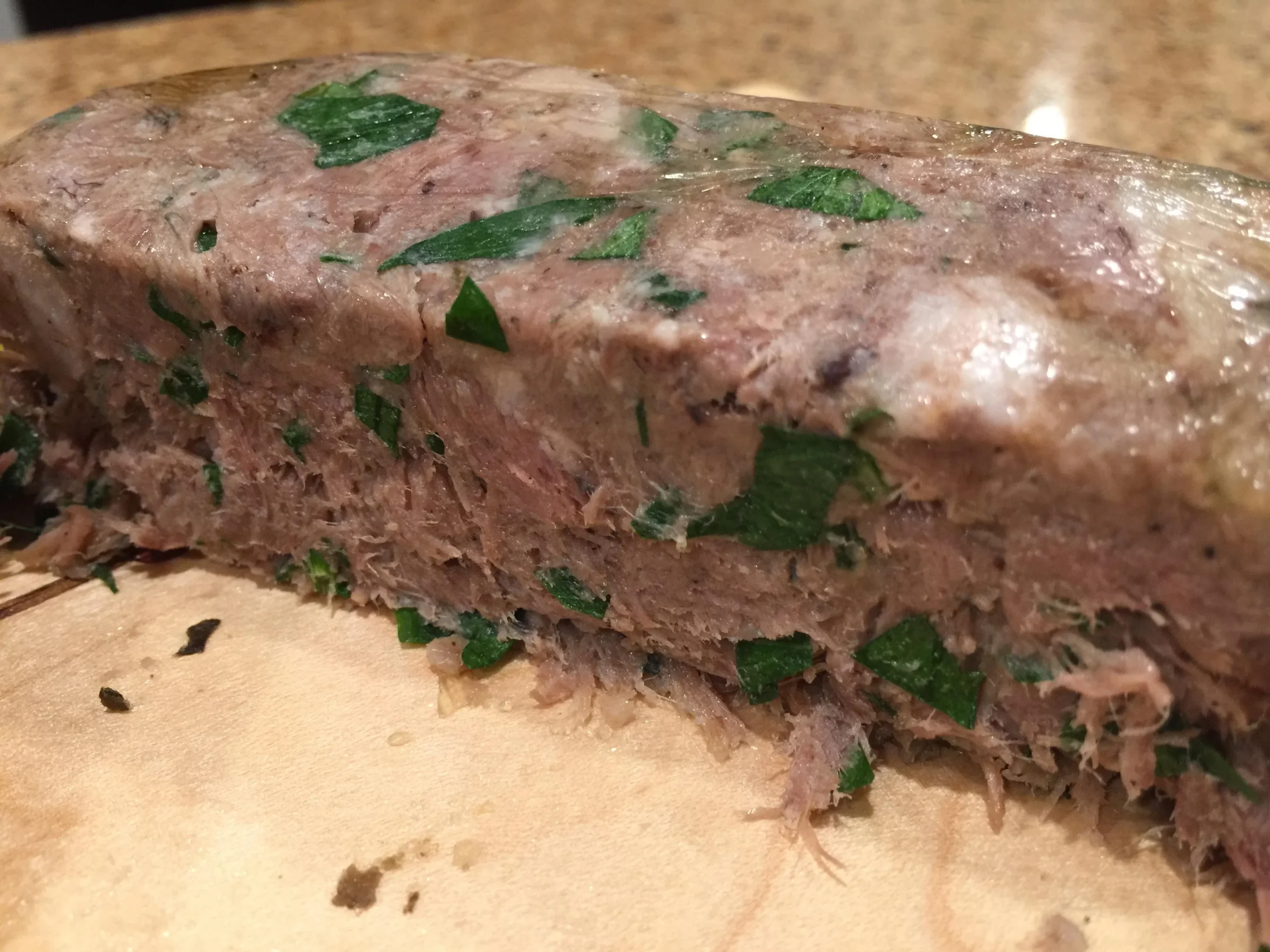 A close-up of a slice of meat terrine with visible herbs, sitting on a wooden cutting board. The mix appears dense and moist, showing green parsley throughout.