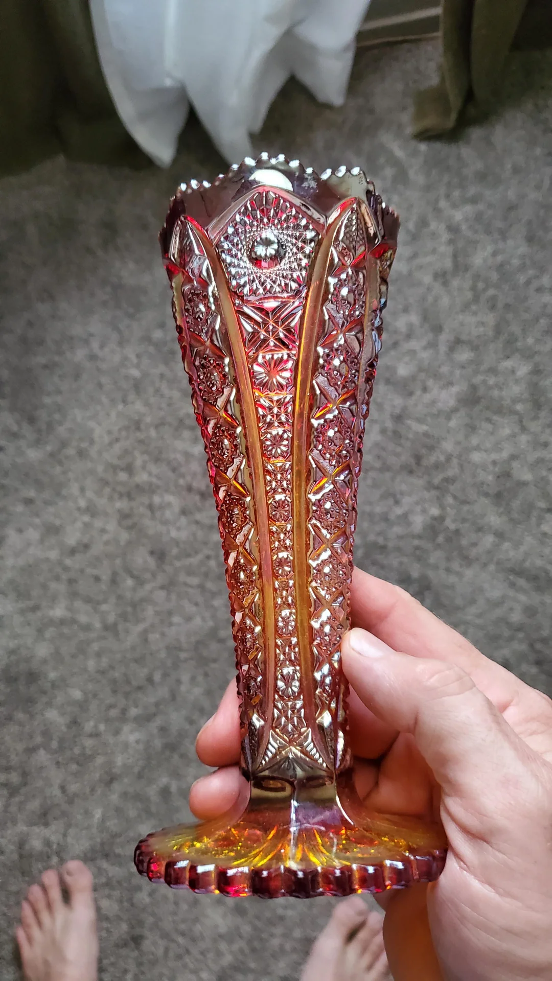A hand holds a tall, ornate, reddish-gold glass vase with intricate patterns and a textured surface against a gray carpet background.