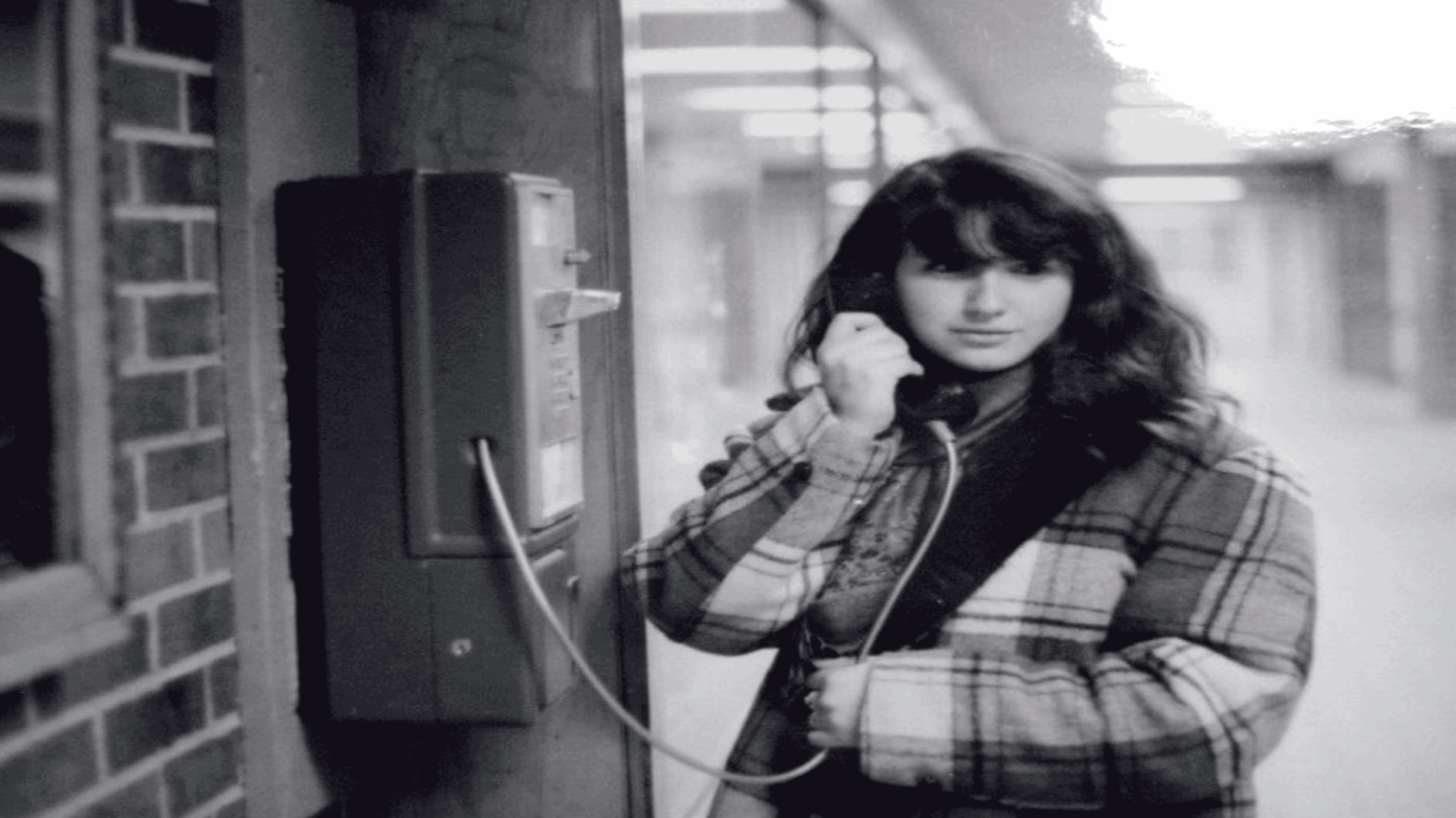 A young woman in a plaid coat uses a payphone in a hallway with brick and glass walls. The image is black and white.