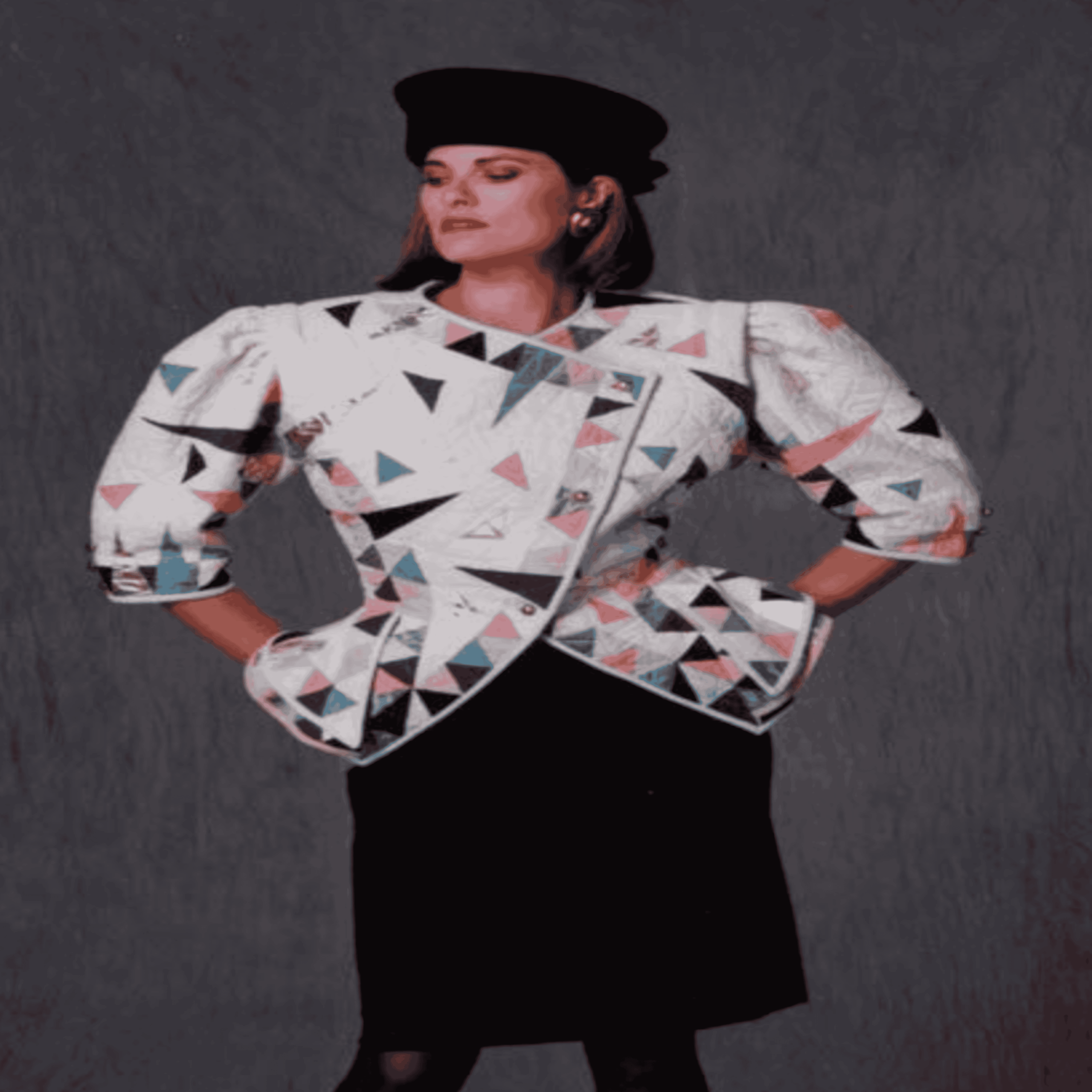 A woman wearing a black hat, black skirt, and a white jacket with colorful geometric triangle patterns poses with her hands on her hips against a dark background.