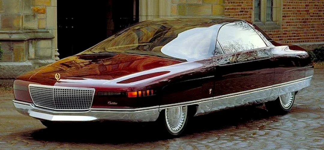 A futuristic, maroon luxury car with a sleek design, wide chrome grille, narrow headlights, and shiny silver trim is parked on a cobblestone street in front of a brick building.