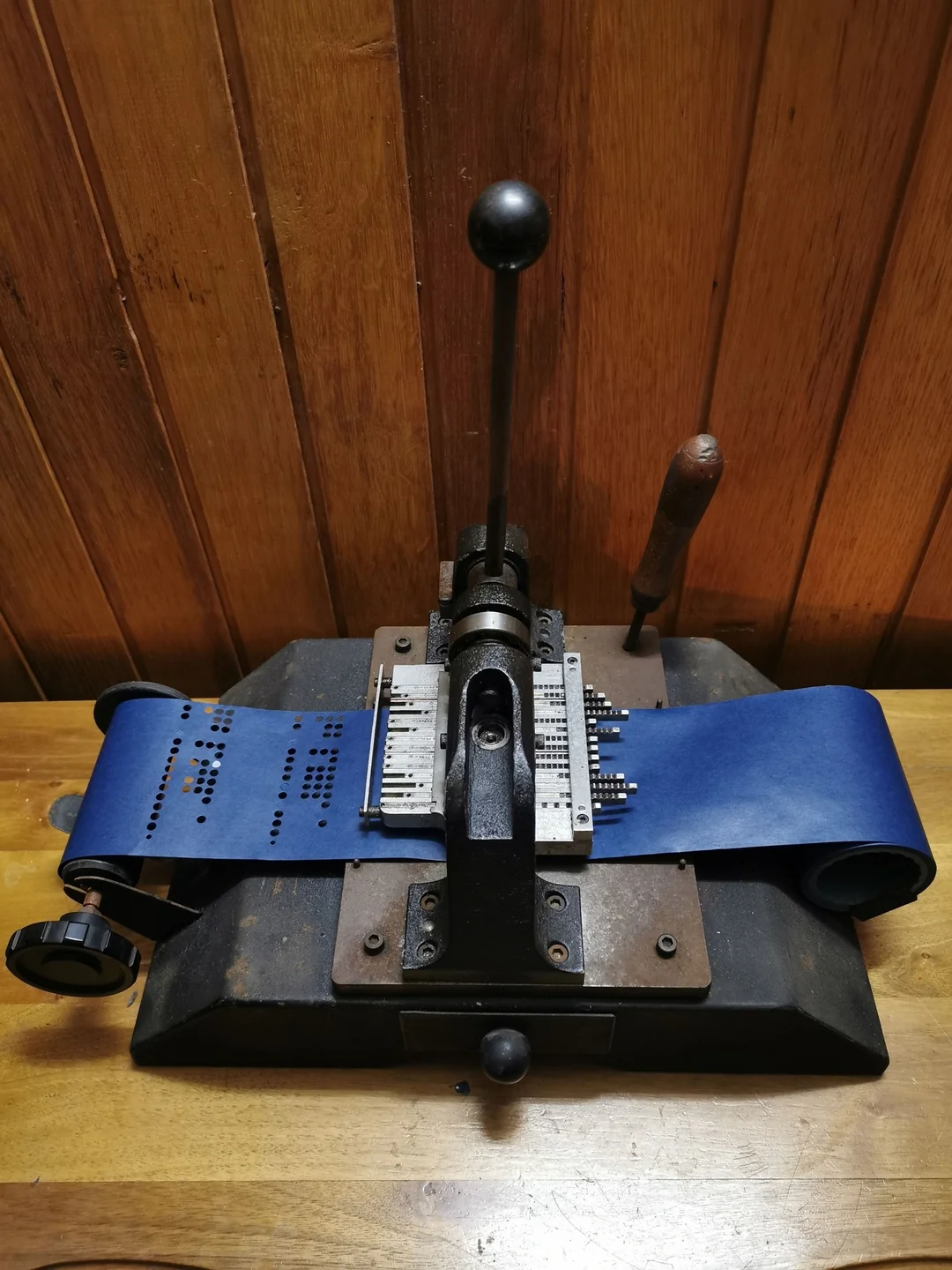 A vintage mechanical punch machine with a handle and a roll of blue paper tape with punched holes, placed on a wooden table against a wood-paneled wall.