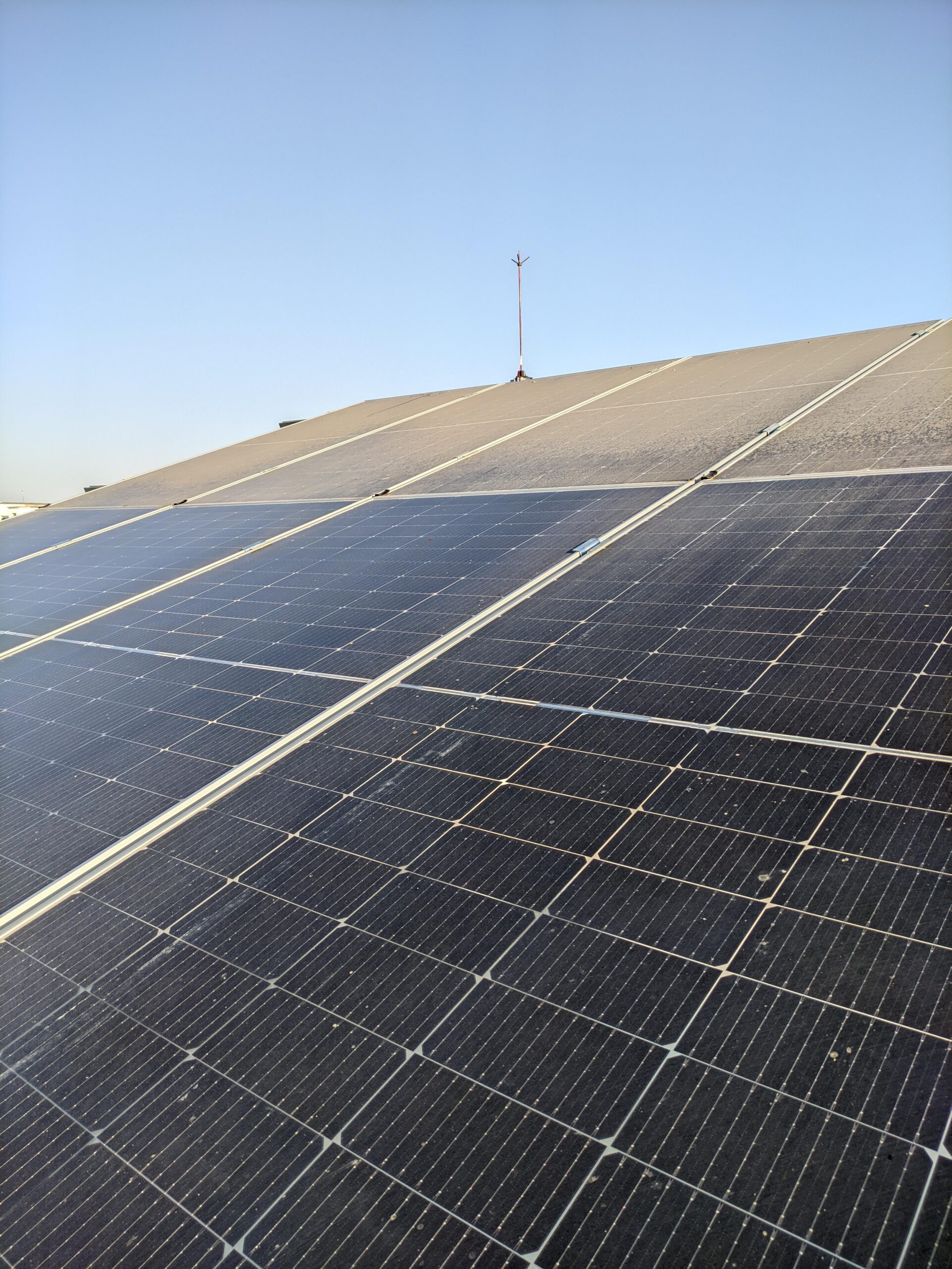 Rows of black solar panels are installed on a slanted rooftop under a clear blue sky, capturing sunlight for energy. A thin antenna rises from the roof’s peak in the background.