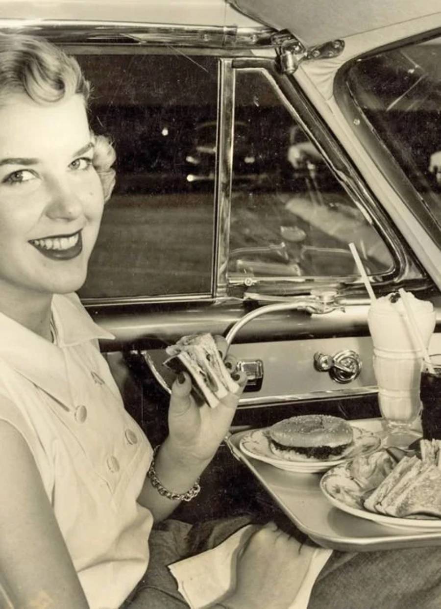 A smiling woman sits in a car, holding a sandwich. On a tray attached to the car window are a burger, fries, and a milkshake. The image is in black and white.