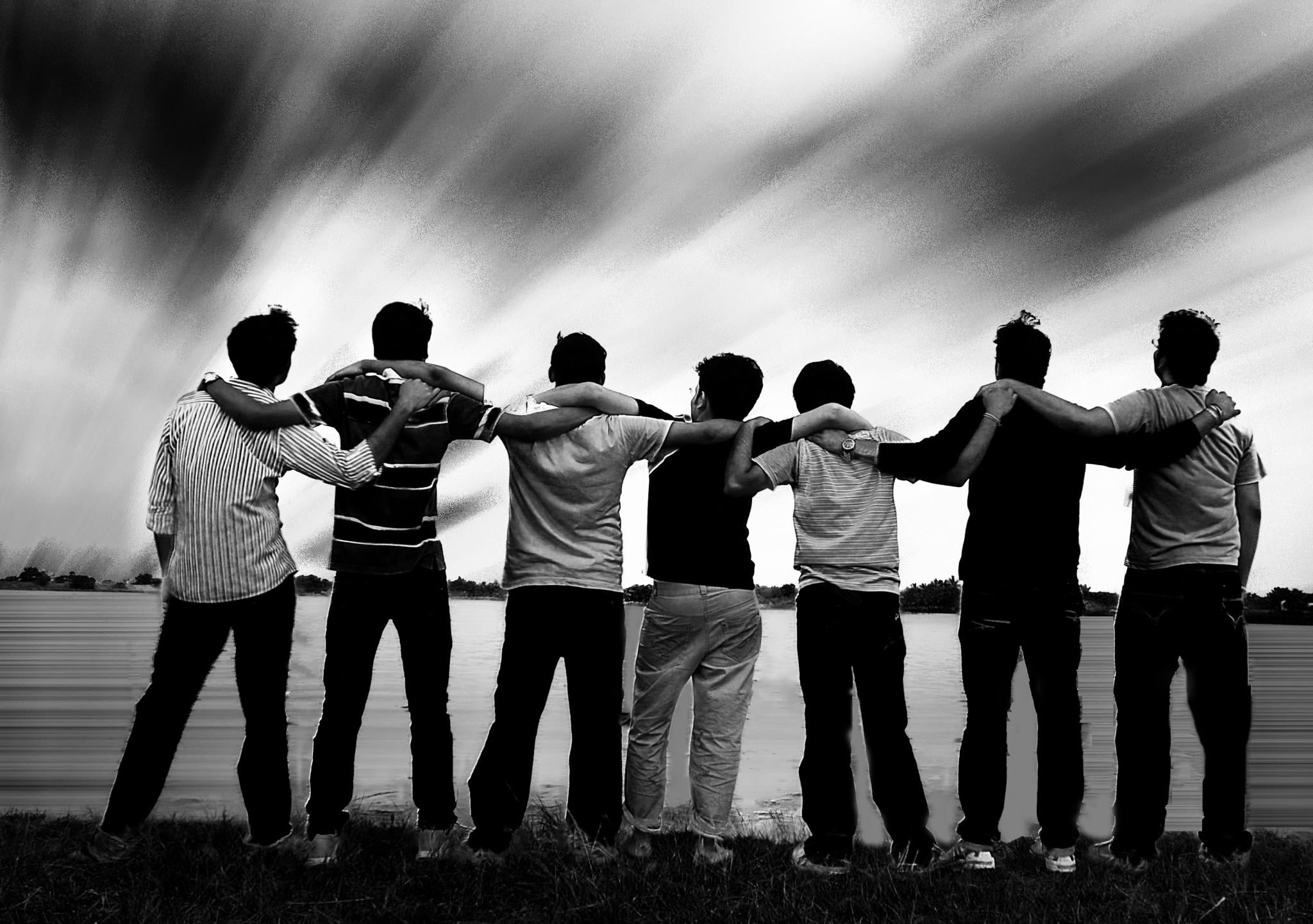 Seven people stand side by side with arms around each other’s shoulders, facing a large body of water under a dramatic, cloudy sky in black and white.