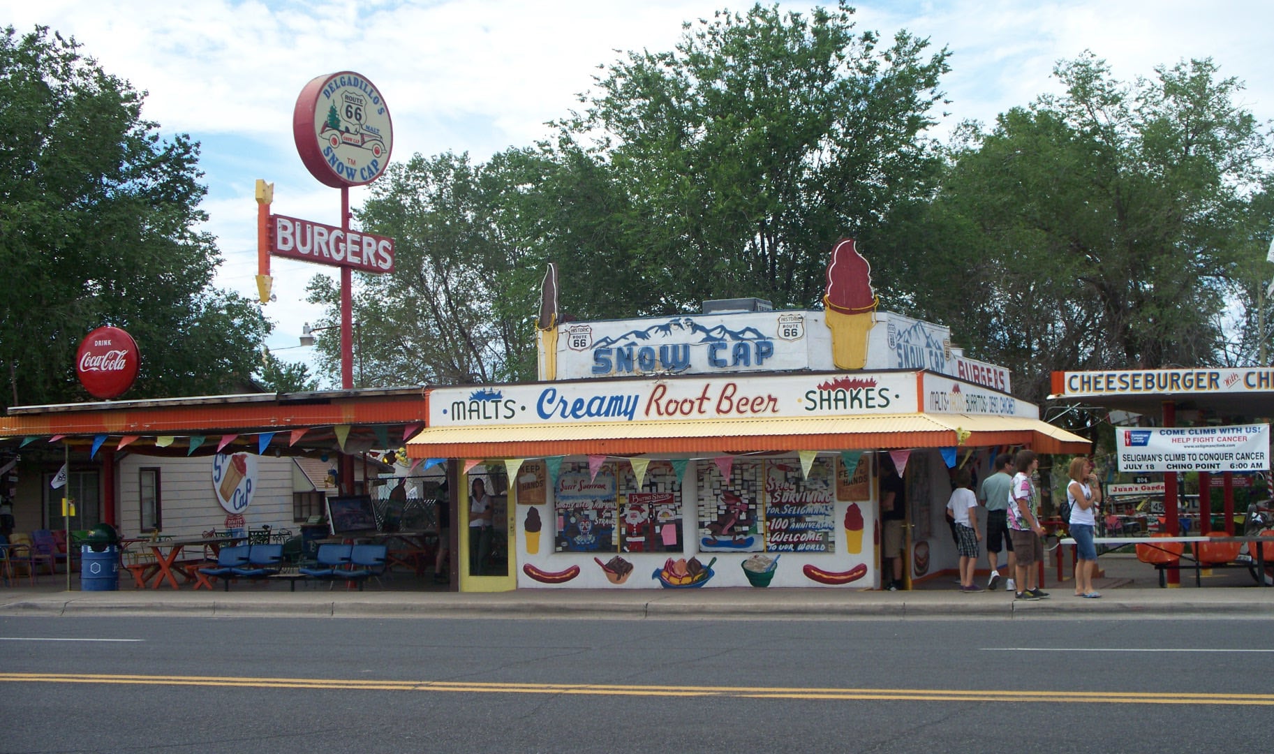 A colorful roadside diner with a sign reading “Snow Cap” offers malts, root beer, and shakes. People are gathered outside. There are vintage burger and Coca-Cola signs, and trees in the background.