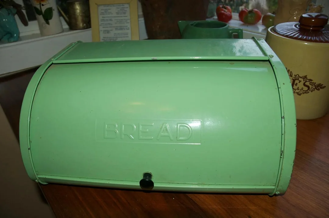 A vintage green metal bread box labeled “BREAD” sits on a wooden countertop, with kitchen items and a window in the background.