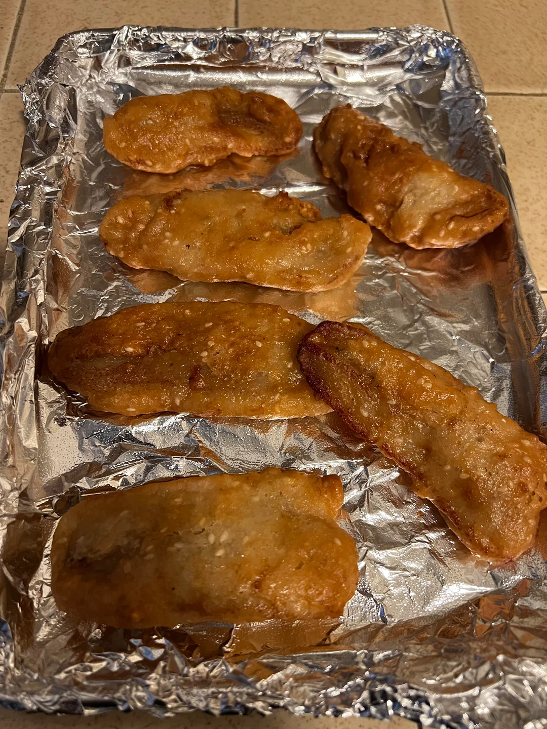 Six pieces of fried food, possibly battered fish or chicken, rest on a foil-lined baking sheet atop a tan countertop.