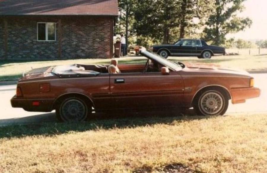 A brown convertible car with its top down is parked on the street near a brick building; another black car is parked in the background.