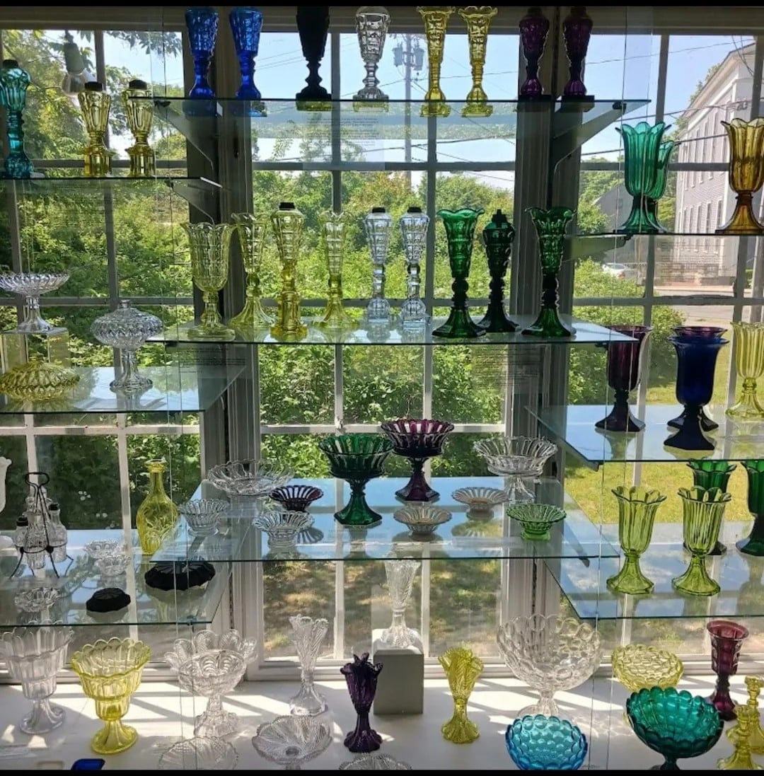 Colorful glass vases and bowls of various shapes are arranged on glass shelves in front of a large window, with trees and a building visible outside.