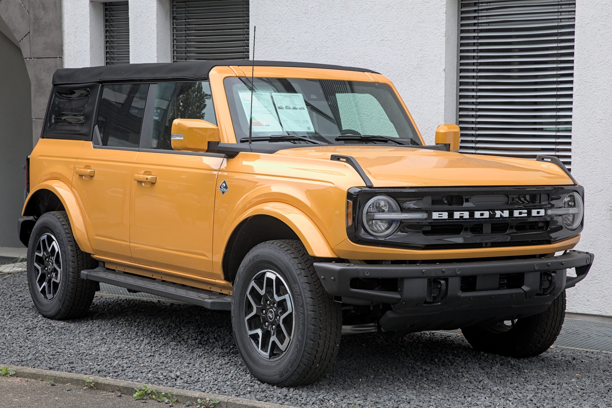 A yellow Ford Bronco SUV with black trim and roof is parked on a gravel area beside a white building with glass windows.