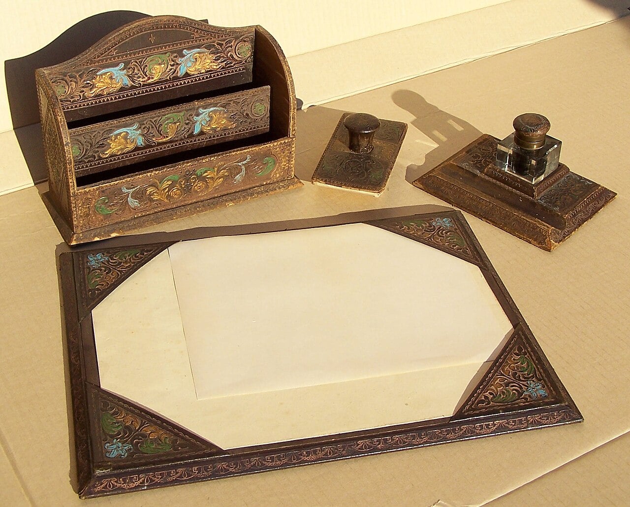 Vintage desk set with ornate brown and gold detailing, including a letter holder, blotter, inkwell, and pen holder, all arranged on a beige surface with a blank sheet of paper.