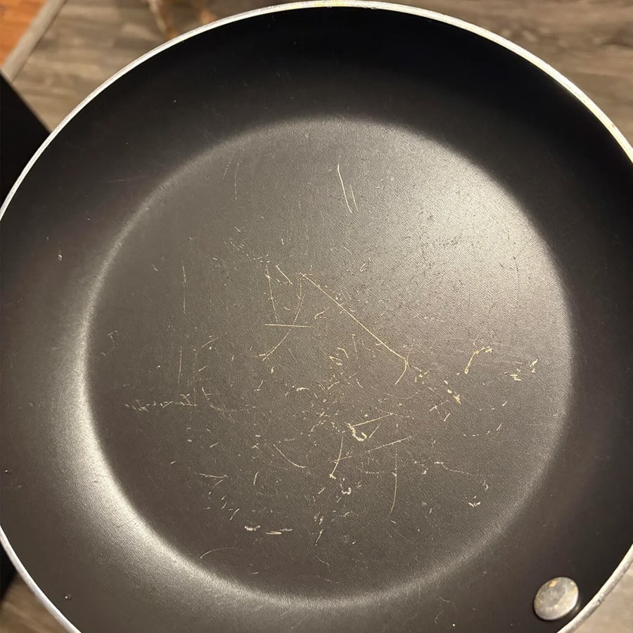 A non-stick frying pan with visible scratches on its surface, indicating wear and tear. The pan is empty and held up against a blurred brown background.