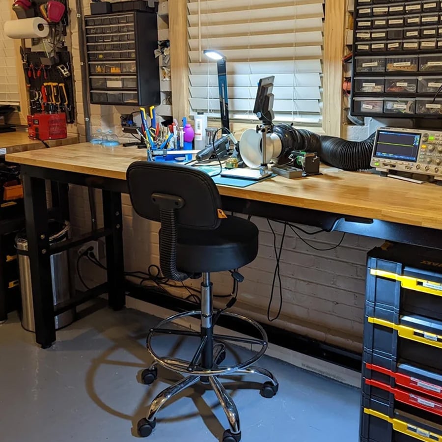 A well-organized electronics workbench with tools, drawers, test equipment, and various supplies. A chair is placed at the wooden workbench under a window with closed blinds.